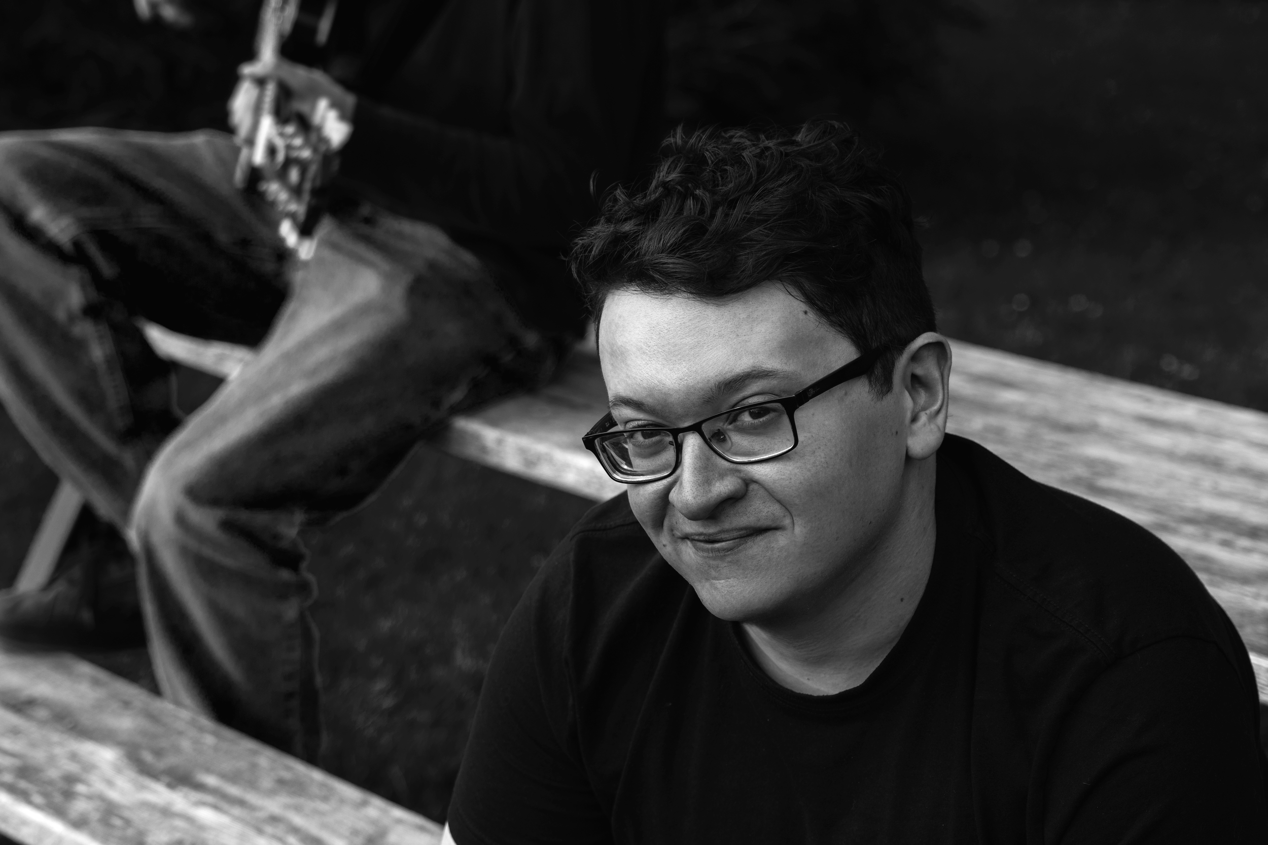 A person with short curly hair and glasses is sitting outdoors at a wooden picnic table, looking at the camera with a slight smile. The background is dark and blurred.