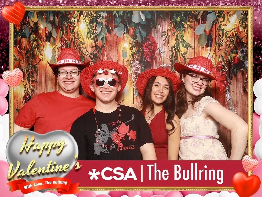 Four young people smiling and posing together at a Valentine's Day themed photo booth, wearing red hats and festive accessories, with a background decorated with flowers, greenery, and string lights.