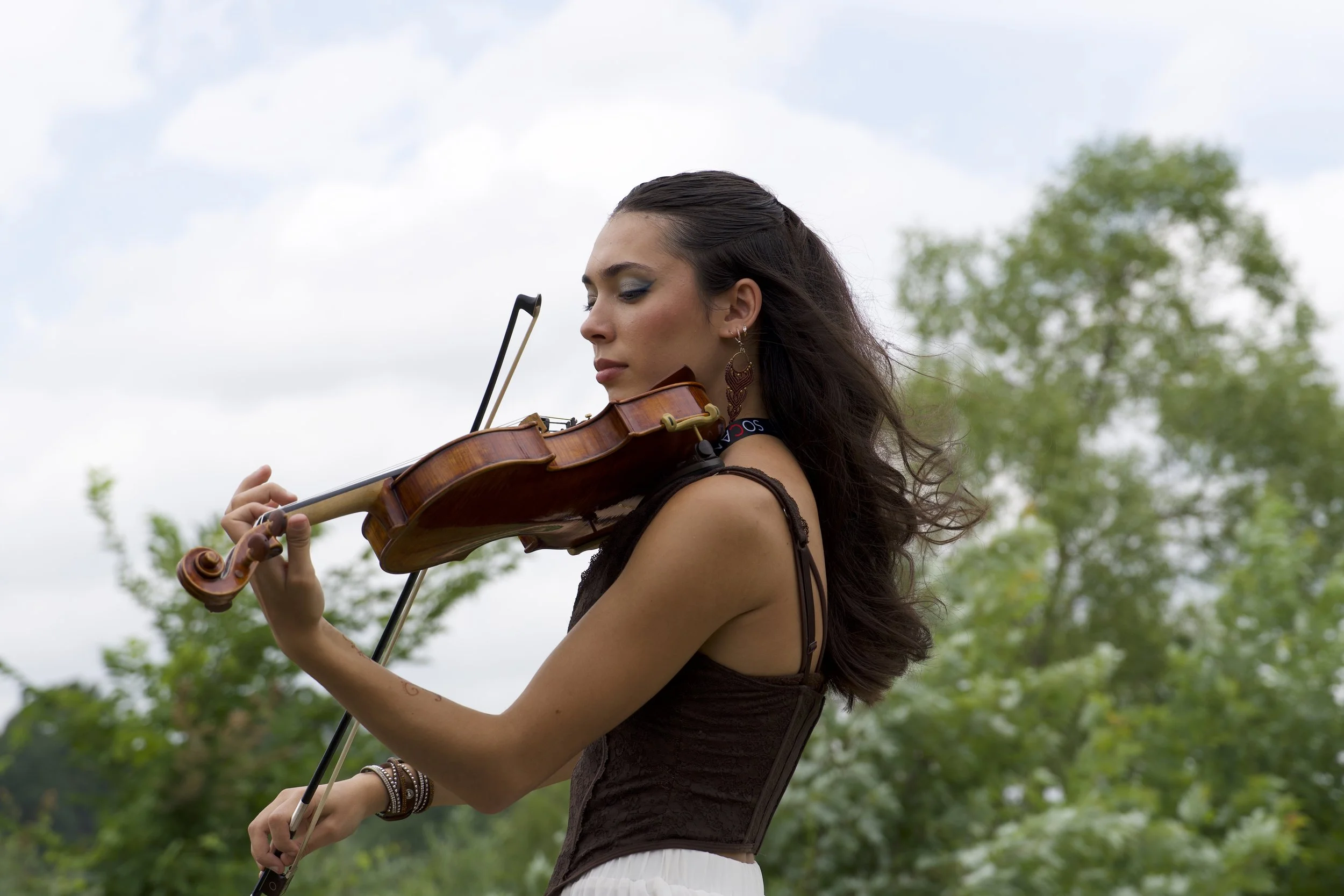 A woman with long dark hair playing the violin outdoors against a cloudy sky with trees in the background.