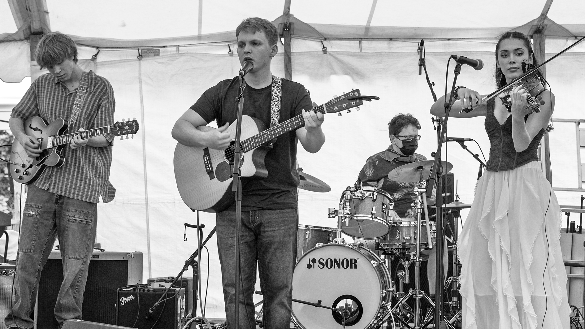 A black and white photo of a four-member band performing on a stage. The band includes a guitarist on the left, a lead singer and guitarist in the middle, a drummer in the background, and a violinist on the right. The musicians are focused on their p