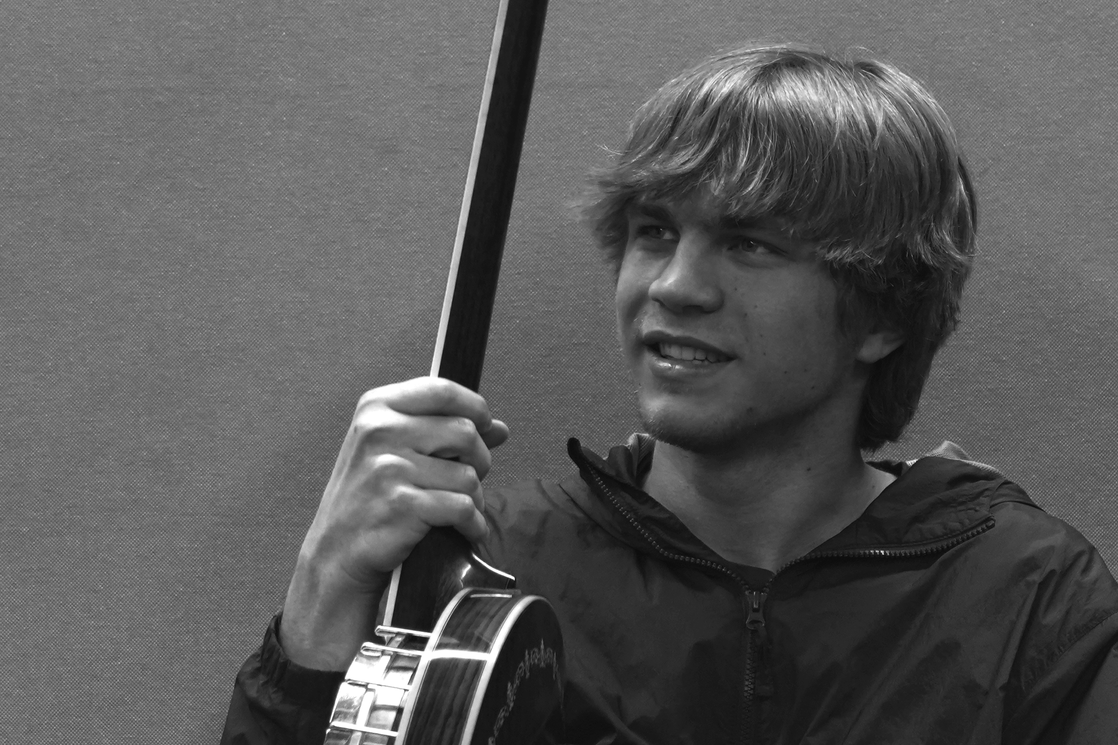 A young man with medium-length hair playing an acoustic guitar and holding a pick, standing in front of a plain textured wall.