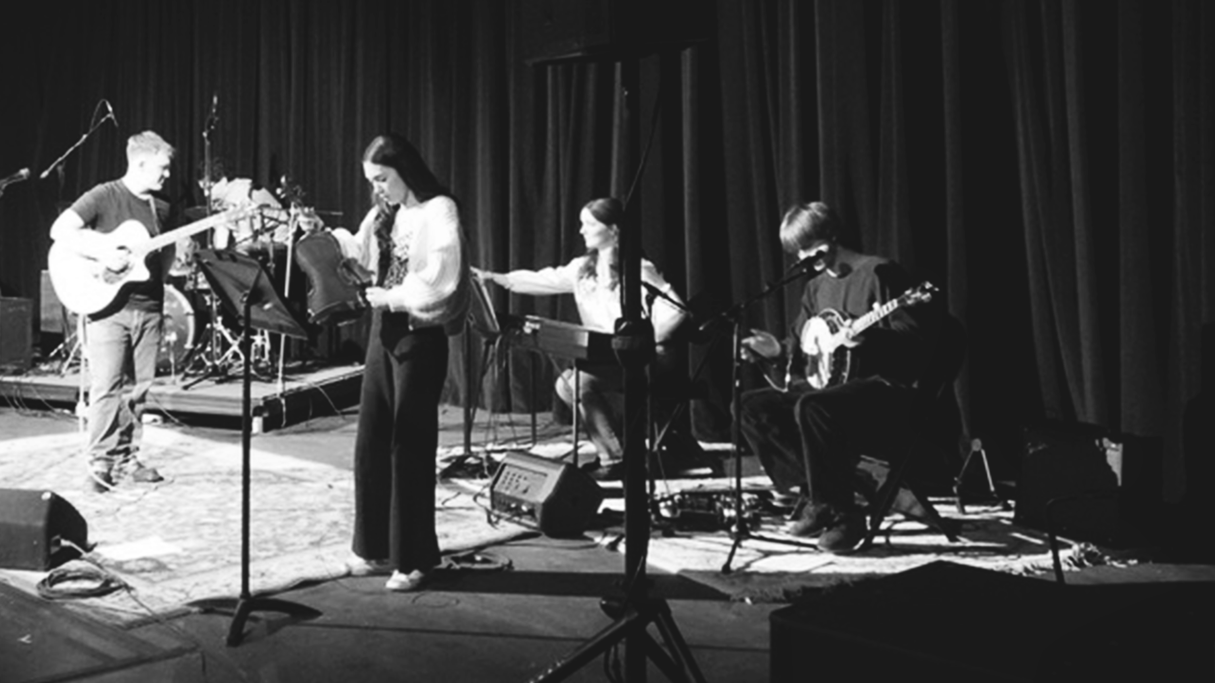 A group of five musicians performing on a stage with dark curtains in the background. One person is playing an acoustic guitar, another is holding a microphone, two others are seated with stringed instruments, and the fifth is adjusting a chair or eq