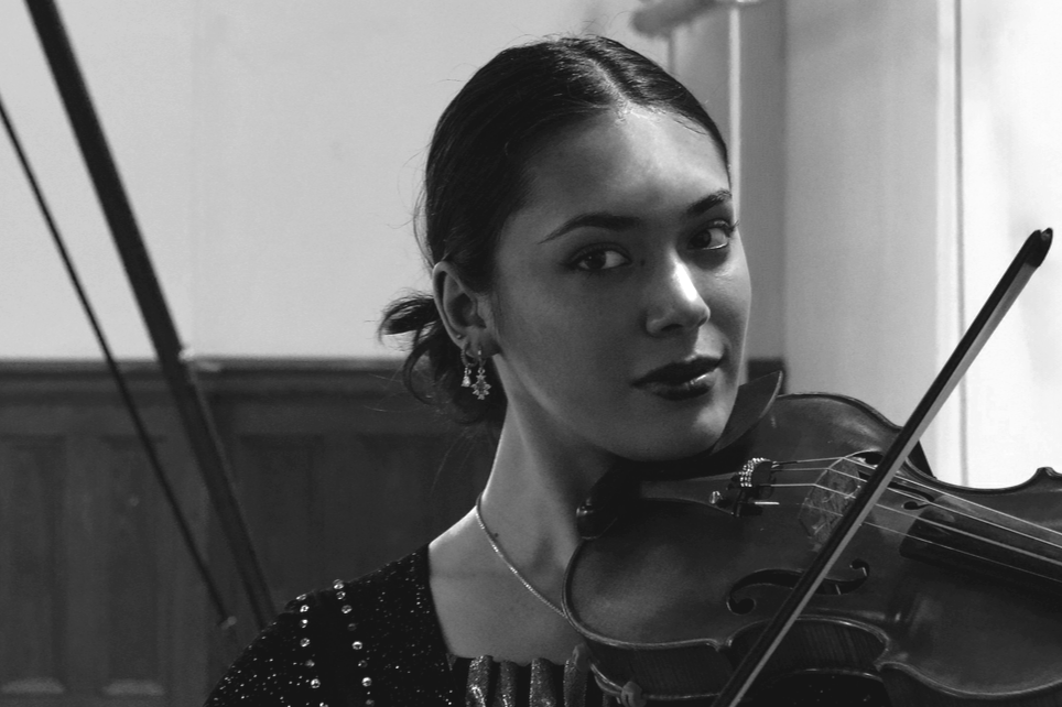 Black and white photo of a woman playing a violin, wearing earrings and a dark dress, with her hair pulled back.