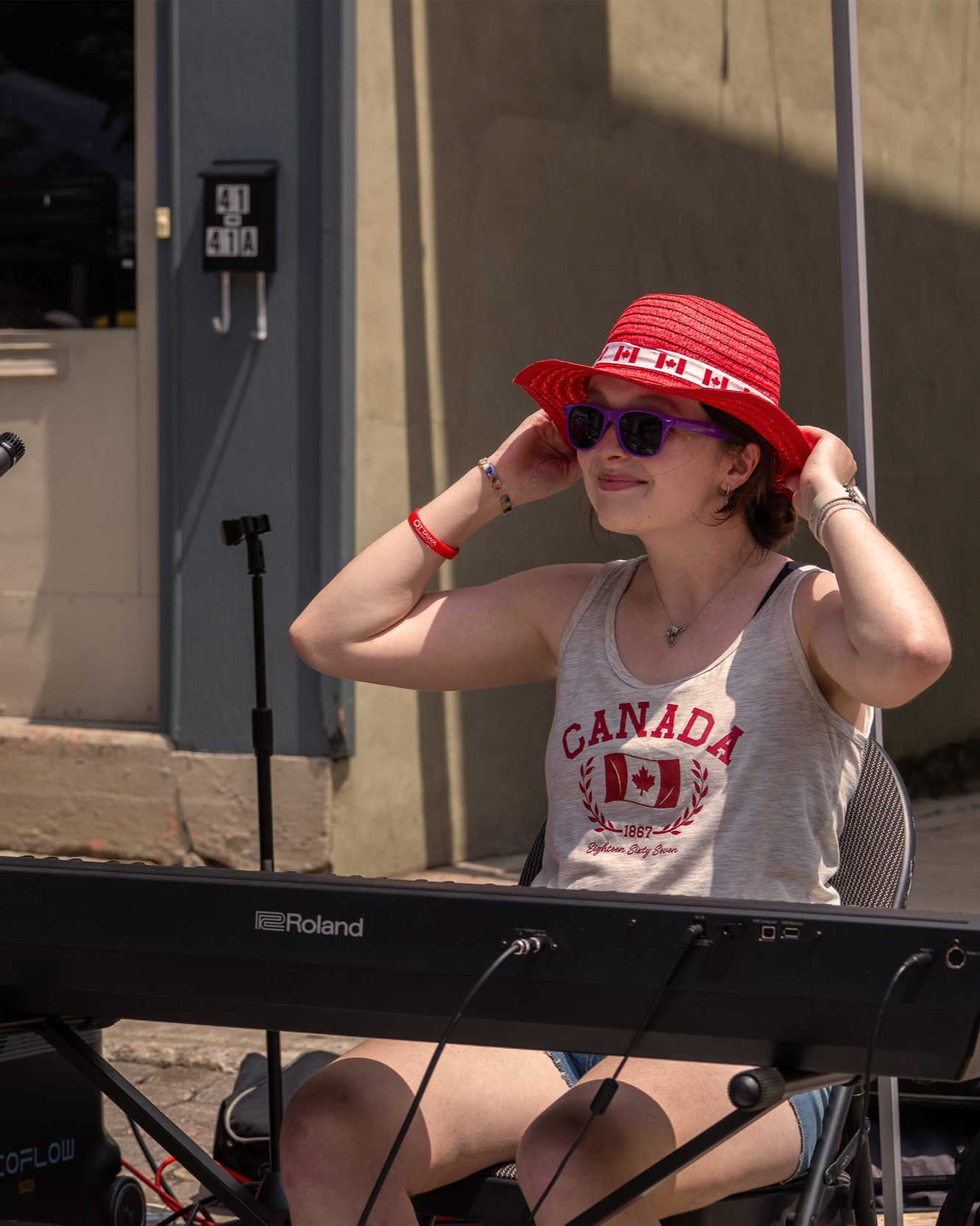 A woman wearing sunglasses, a red sun hat with Canadian flag design, and a sleeveless gray shirt with Canada emblem, sitting at a keyboard outdoors on a sunny day.