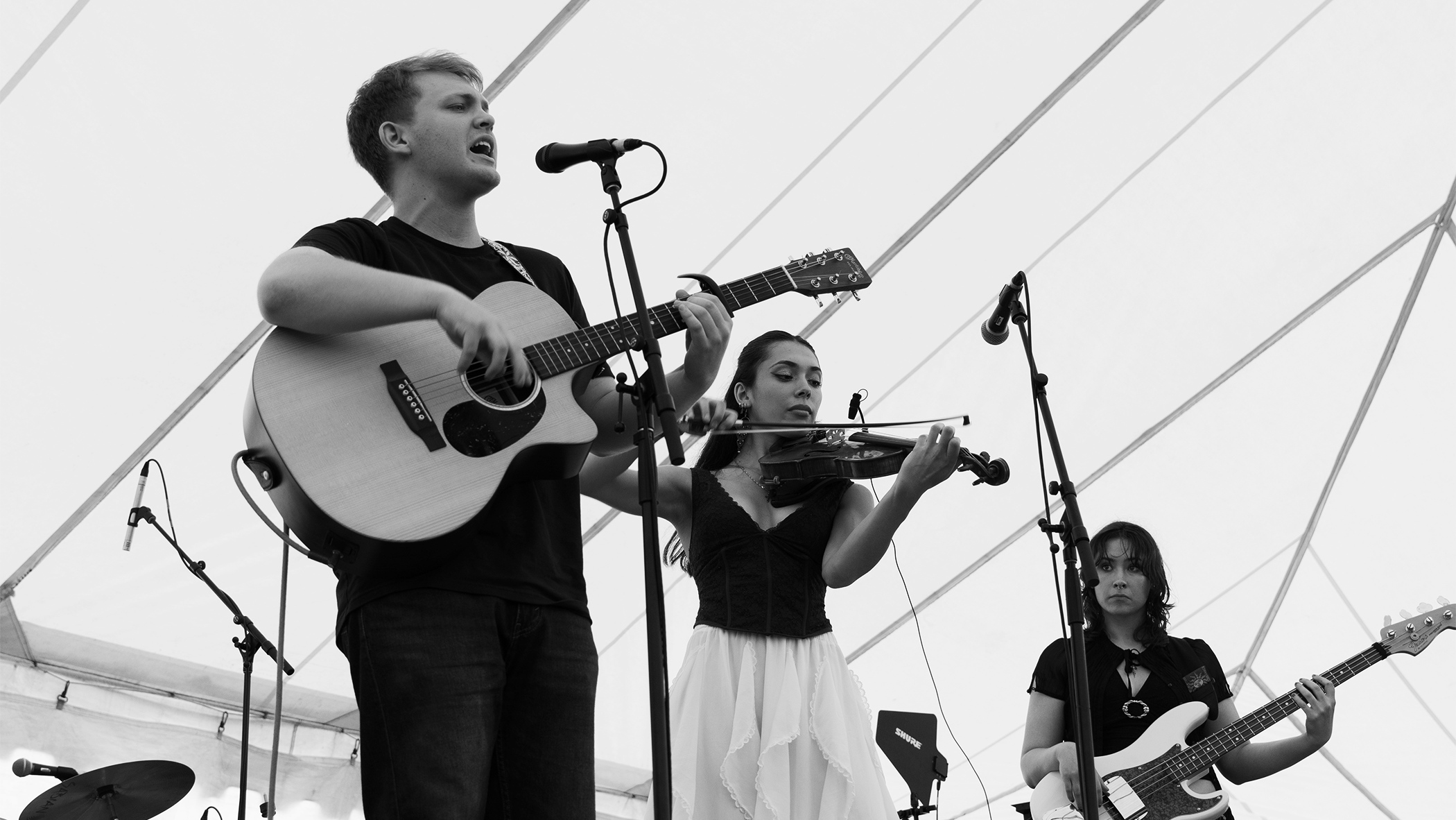A black and white photo of a band performing on stage, featuring a male guitarist and singer, a female violinist, and a female bass guitarist.