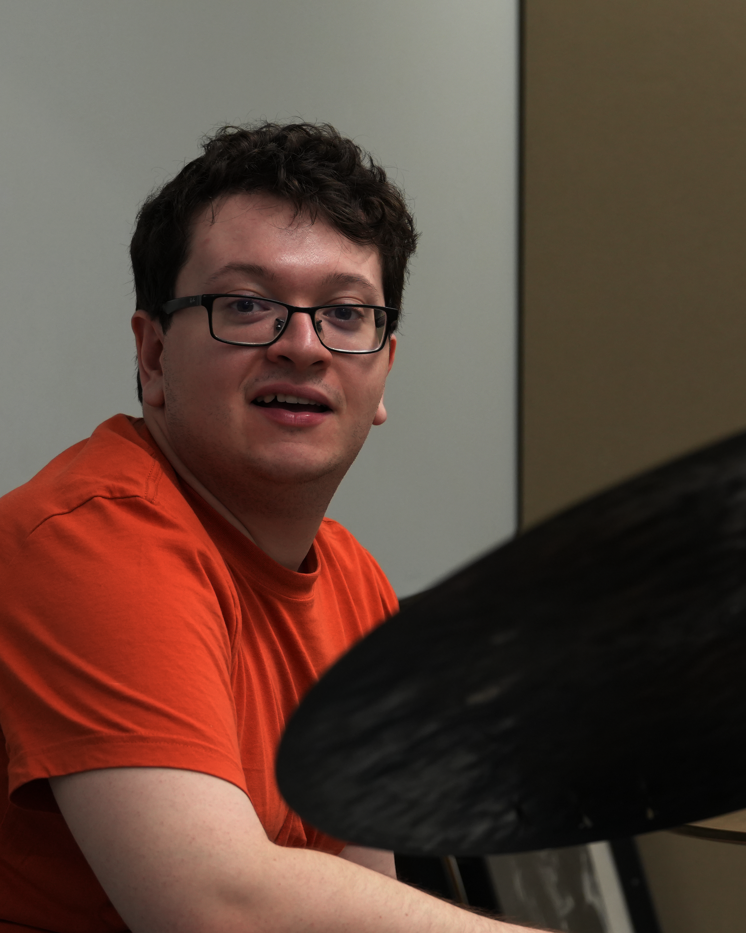 A young man with curly brown hair, glasses, and fair skin, wearing an orange T-shirt, appears to be speaking or singing, with his mouth slightly open, in an indoor setting with plain and dark walls.