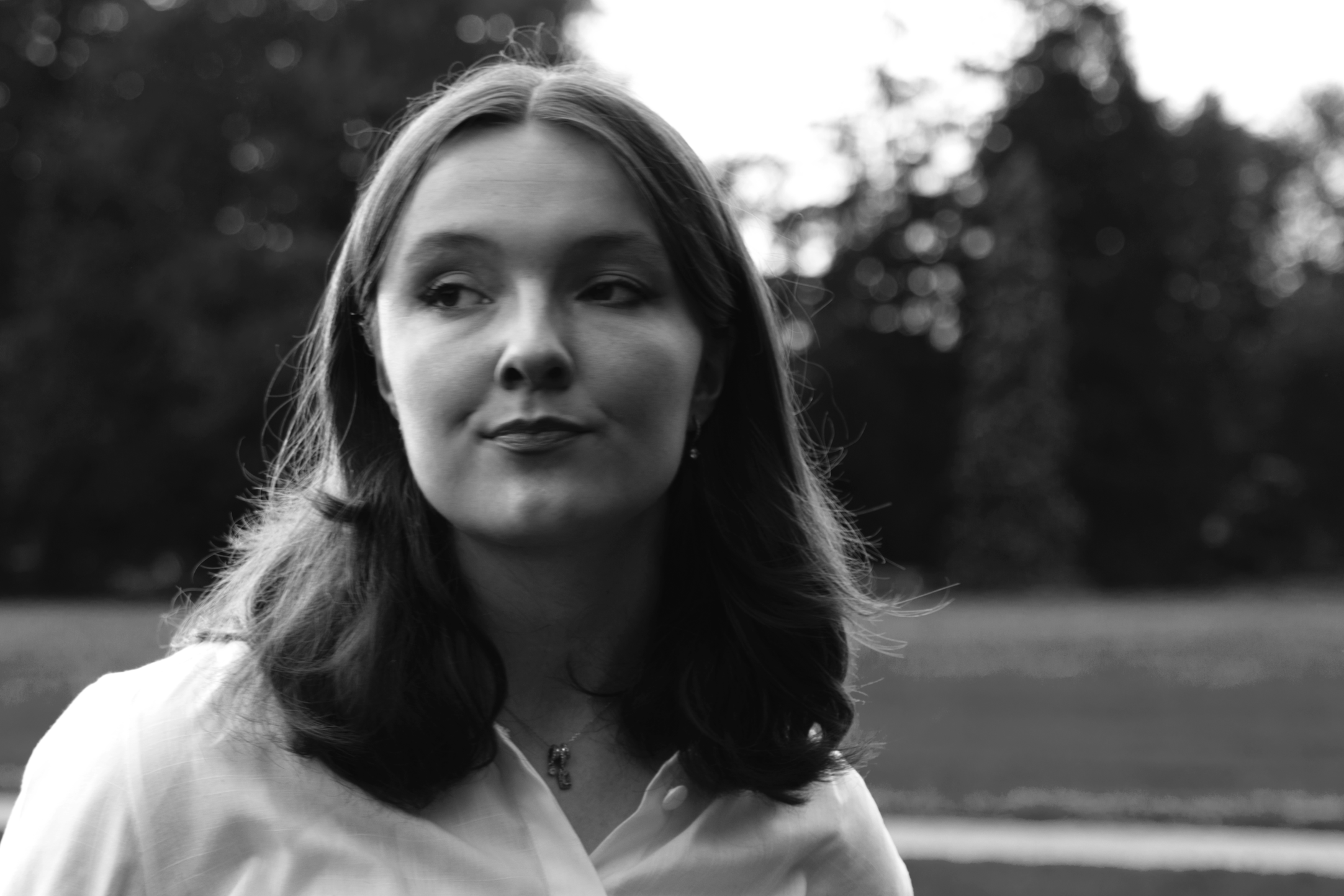Black and white portrait of a woman with medium length hair outdoors, with blurred trees in the background.