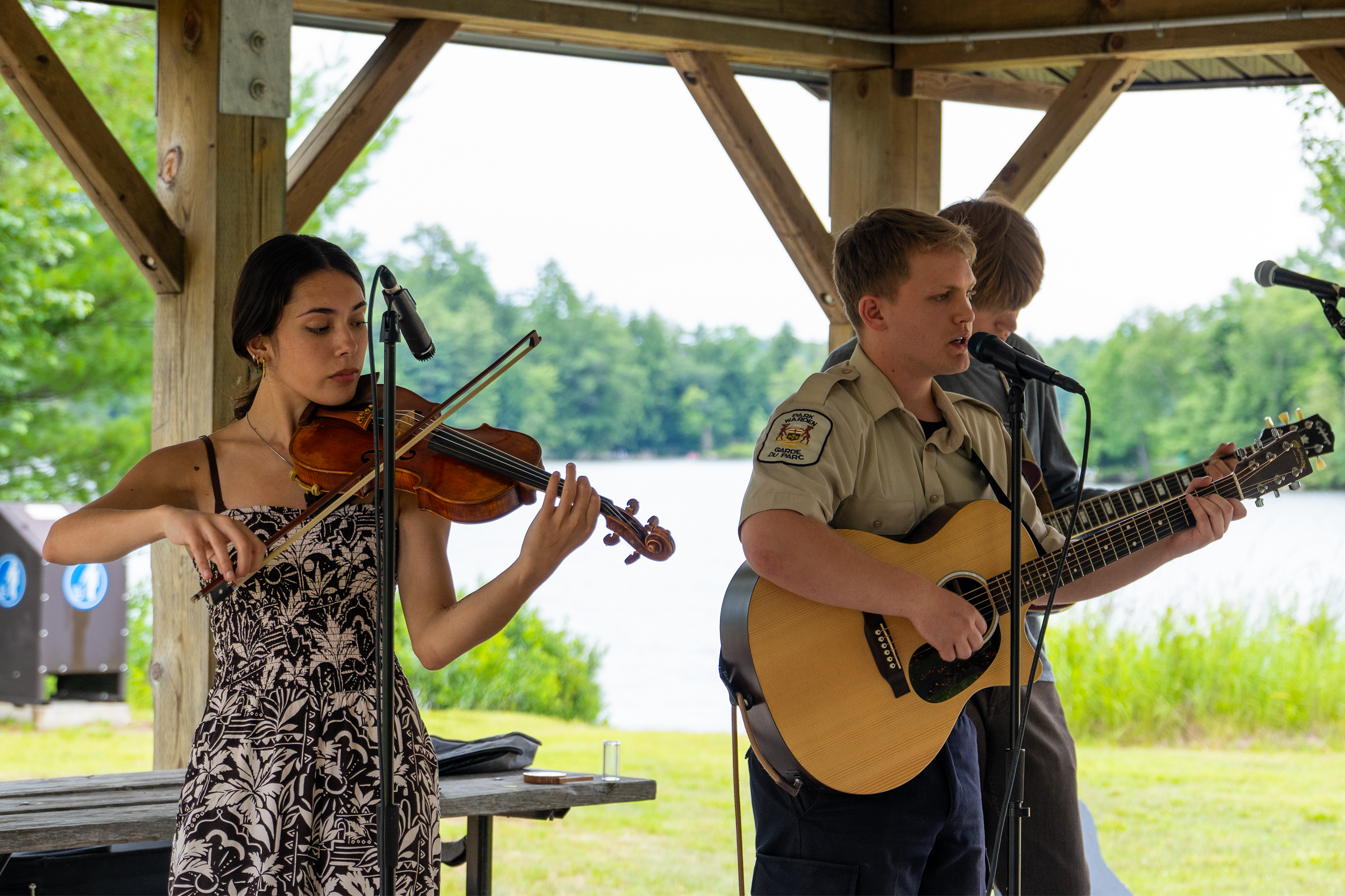 A young woman playing a violin and a young man singing and playing an acoustic guitar outdoors under a wooden pavilion.