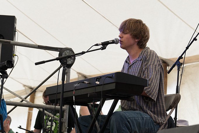 Young man playing a keyboard and singing into a microphone under a tent at an outdoor event.