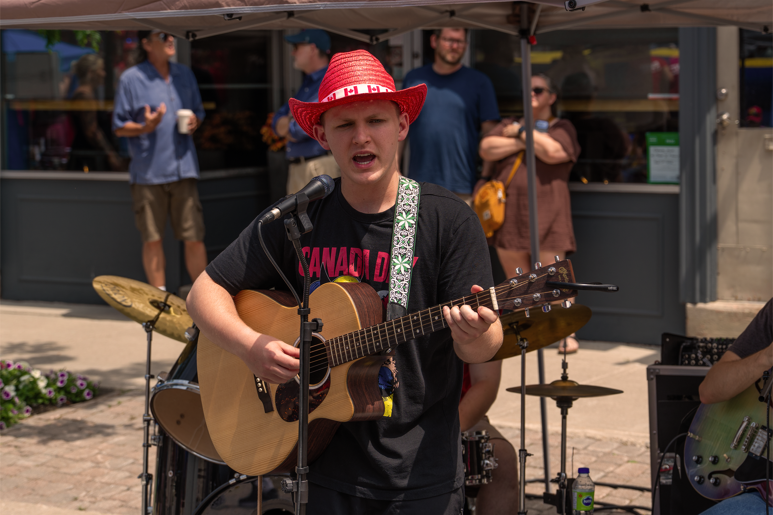 Young man in a red patriotic hat playing an acoustic guitar and singing into a microphone during an outdoor performance, with people in the background.