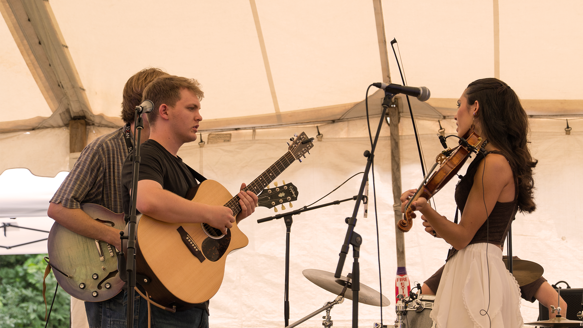 Two musicians, a young man and a young woman, playing guitars and a violin during a performance inside a tent, with microphones set up in front of them.