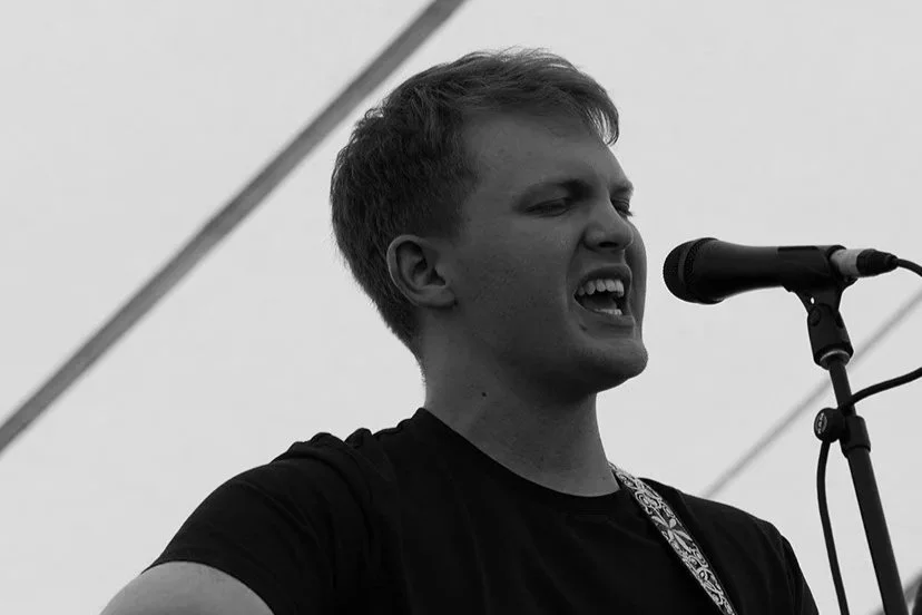 Young man singing into a microphone, wearing a black shirt, with a guitar strap over his shoulder, performing on stage.