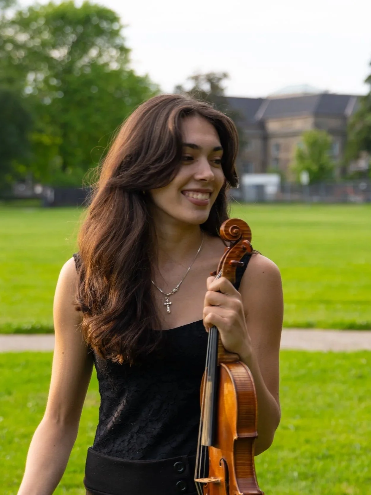 A young woman smiling and holding a violin outdoors in a park with green grass and trees in the background.