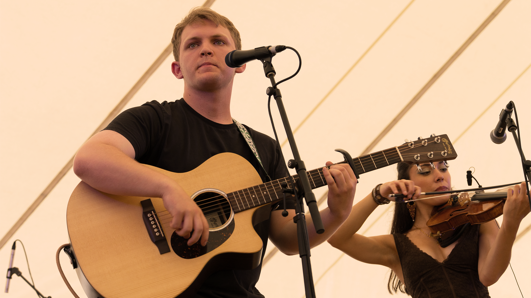 A young man playing an acoustic guitar and singing into a microphone, accompanied by a young woman playing a violin. They are performing under a large, white tent or canopy.