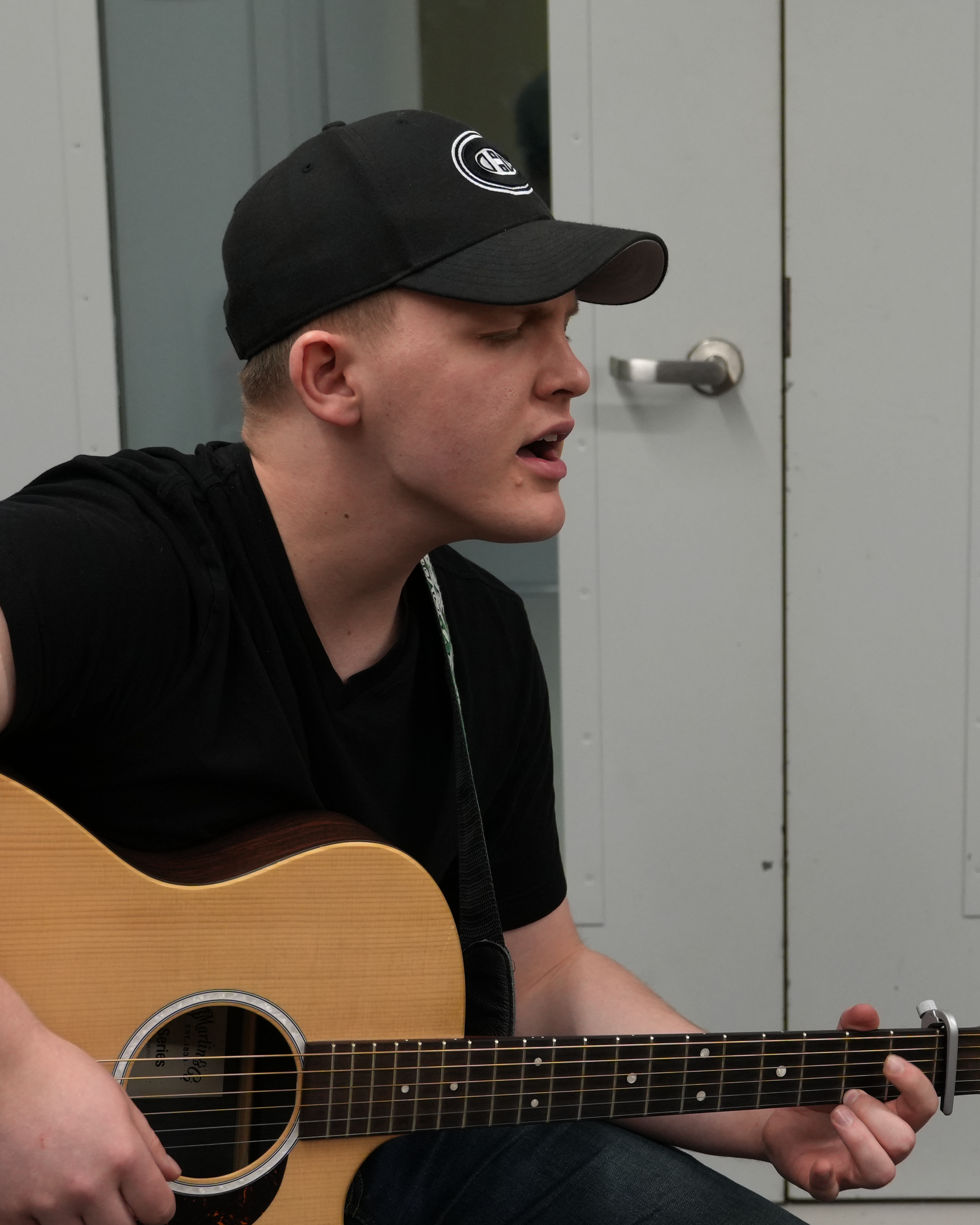 A young man is playing an acoustic guitar while sitting down. He is wearing a black baseball cap with a logo and a black shirt. Behind him, there is a white door with a silver handle.