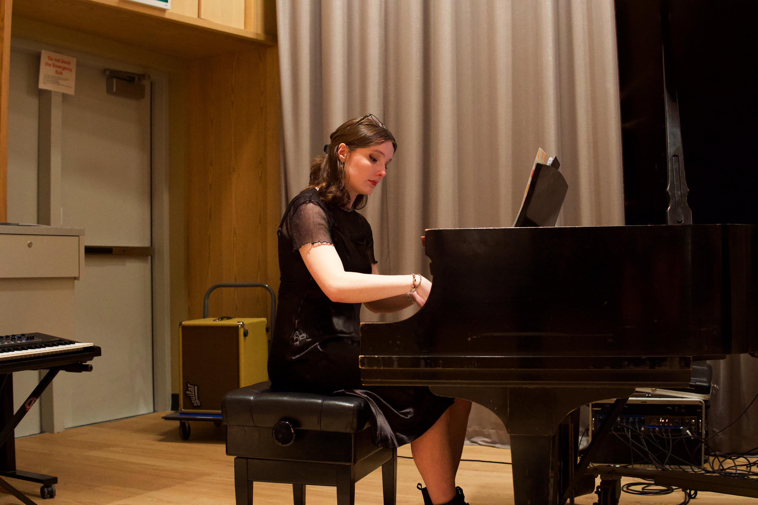 A young woman with shoulder-length brown hair wearing a black dress sitting at a grand piano, playing it in a room with wooden walls, curtains, and musical equipment.