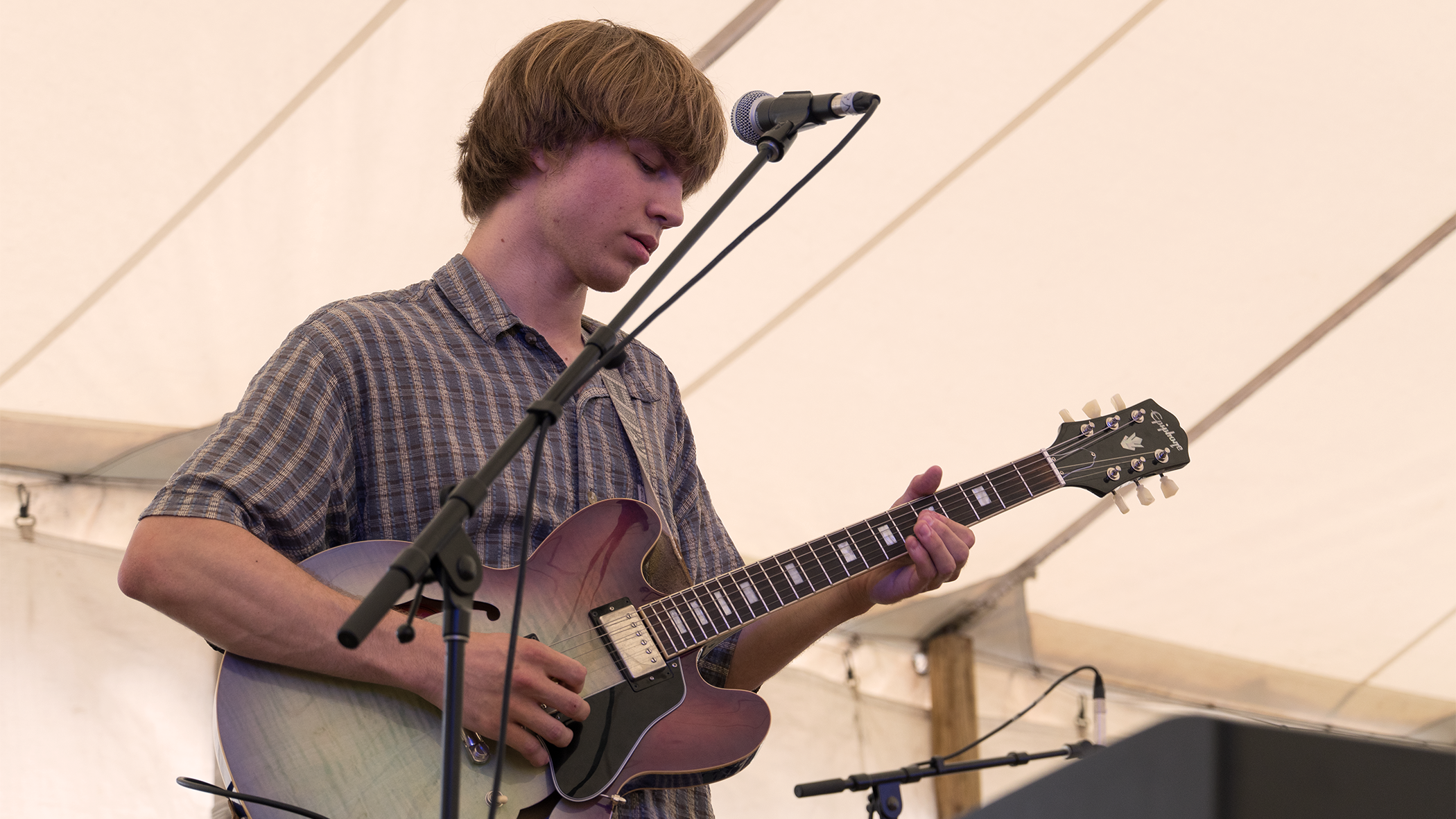 A young man with brown hair and a plaid shirt playing an electric guitar on stage under a tent.