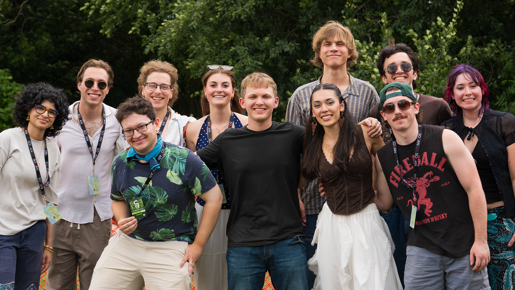 Group of diverse young adults smiling outdoors in front of green trees, some wearing casual clothes, sunglasses, and lanyards.