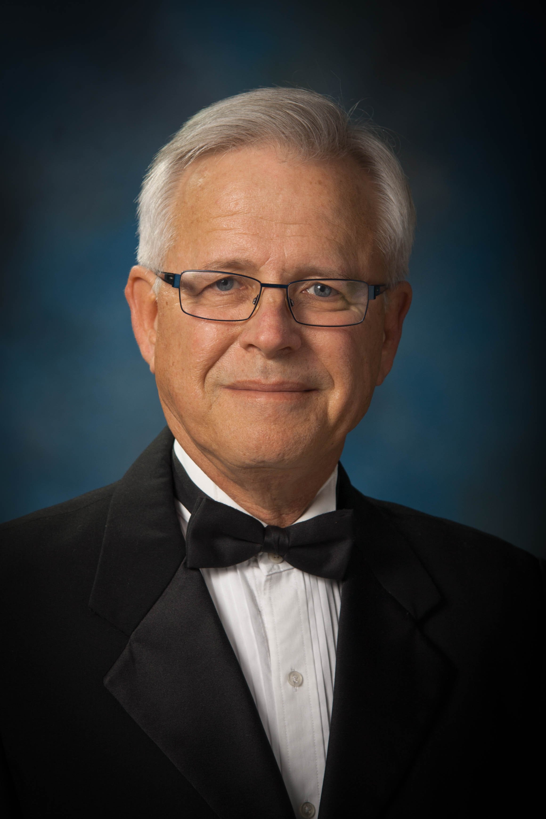 A senior man wearing glasses and a tuxedo with a bow tie, smiling on a blue gradient background.