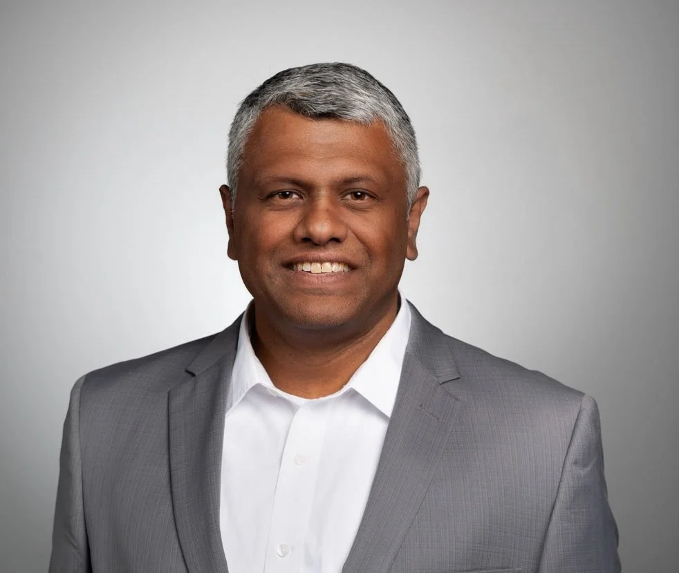 Professional portrait of a smiling man with short gray hair, wearing a light gray suit and white shirt, against a plain gray background.