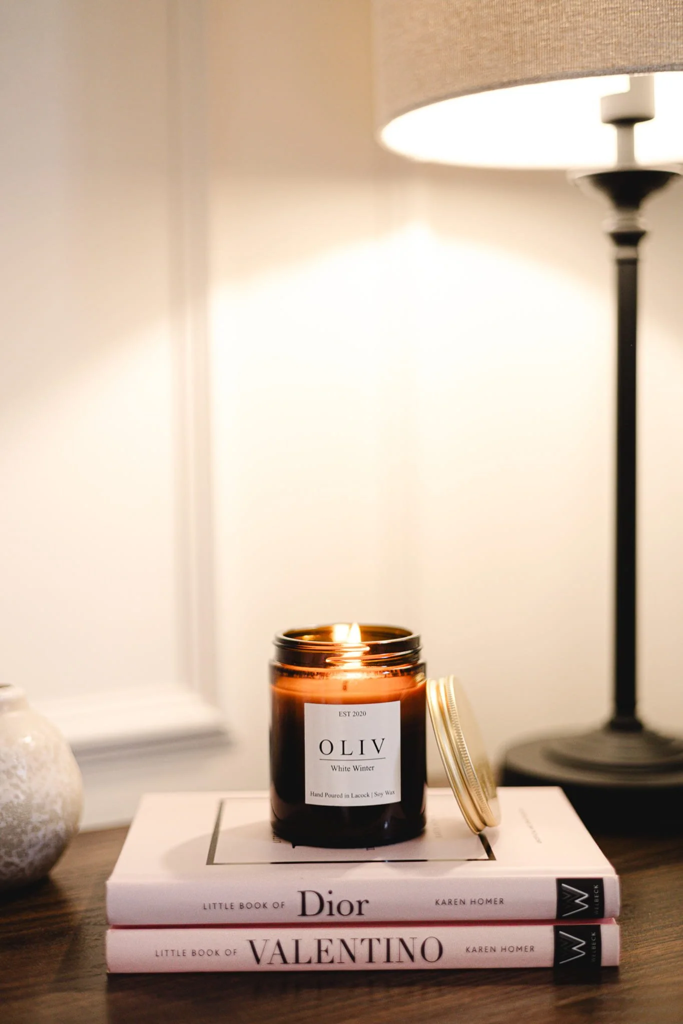 Decorative scene on a wooden surface with two hardcover fashion books, a lit candle in a dark amber jar, a small white textured vase, and a tall black table lamp with a beige lampshade.