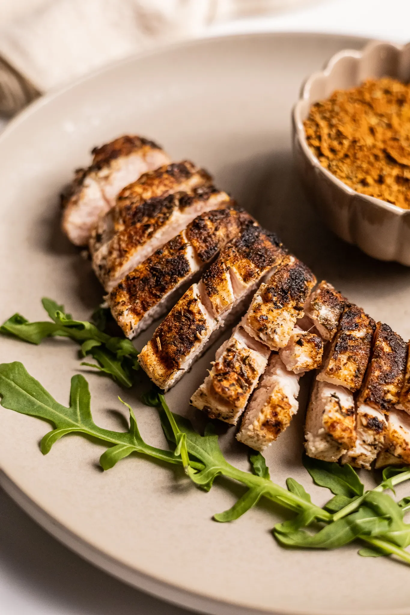 Sliced grilled chicken breast with seasoning, garnished with arugula leaves, served on a beige plate with a side of spicy seasoning or sauce in a small bowl.