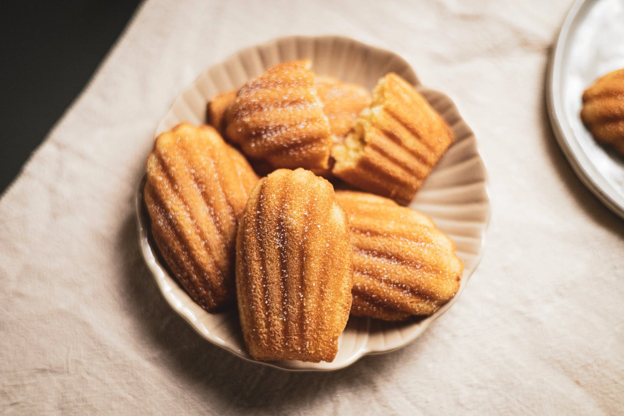Six golden-brown madeleines in a shell-shaped dish on a light-colored cloth surface.