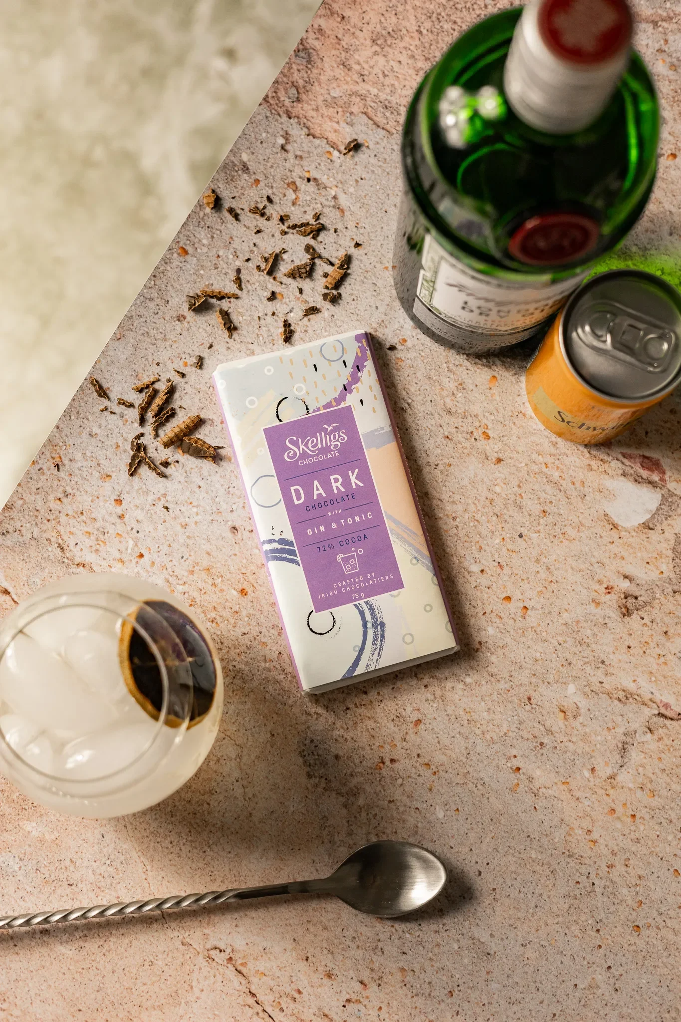 A bar scene with a Skelligs Dark Chocolate bar, a glass of beer, a wine bottle, a beer can, and scattered chocolate shavings on a textured surface.