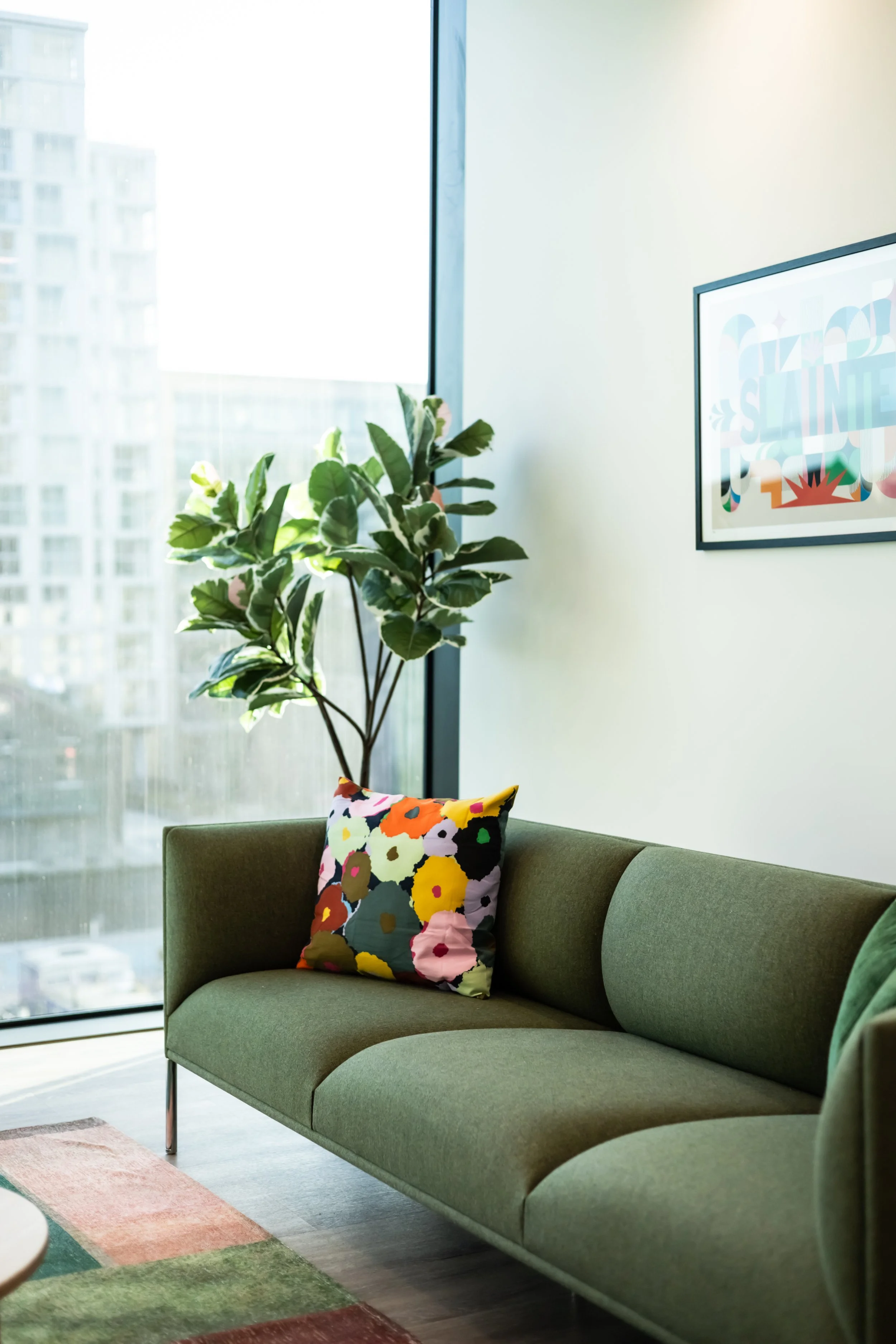 A modern living room corner with a green sofa, a colorful floral cushion, a large leafy plant, and a framed colorful artwork on the wall. Sunlight streams through a large window showing a cityscape outside.