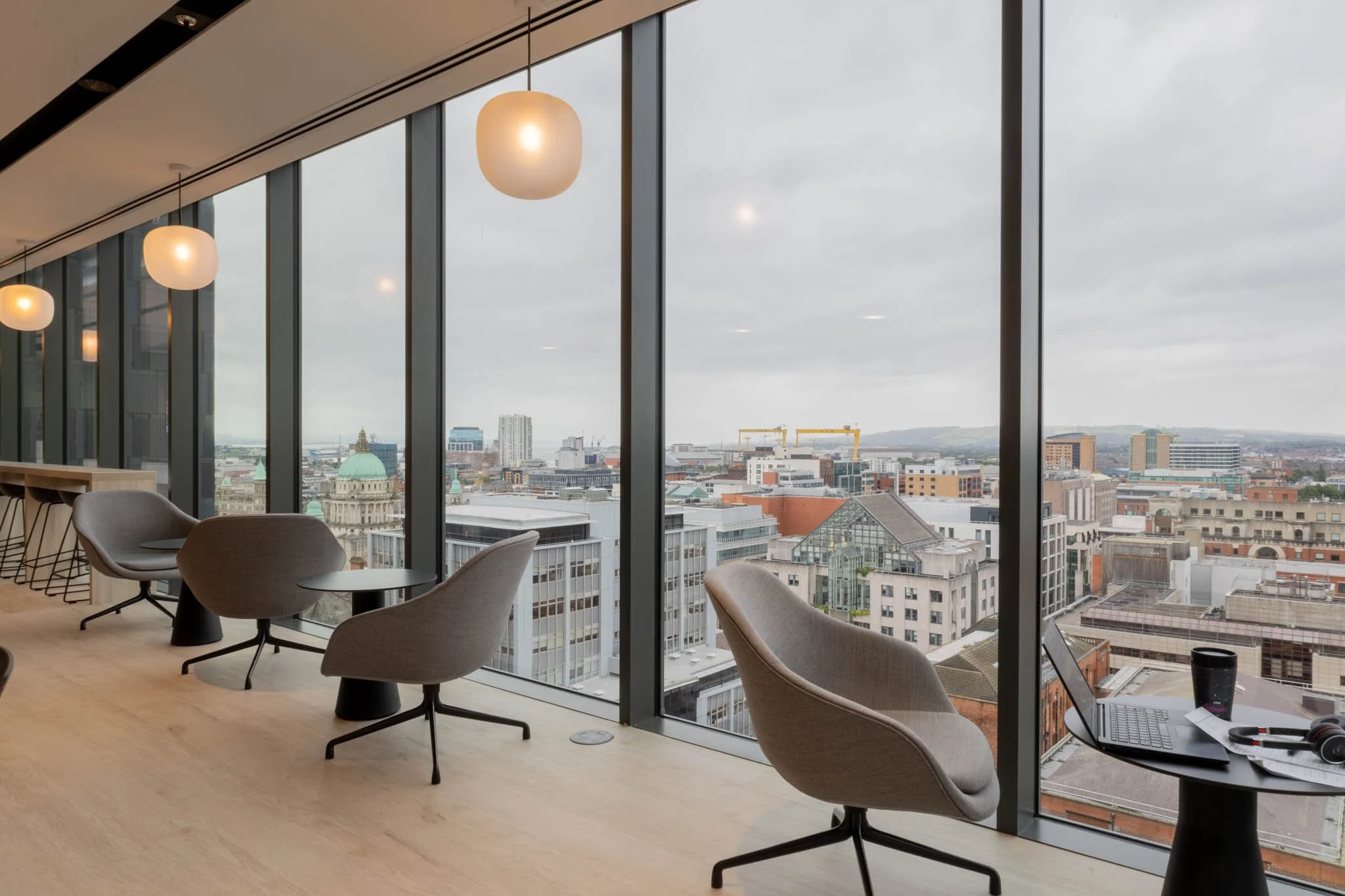 Modern office with large floor-to-ceiling windows offering a cityscape view, interior chairs, small tables, hanging light fixtures, and a laptop with headphones and a coffee cup on a table captured for architectural photography