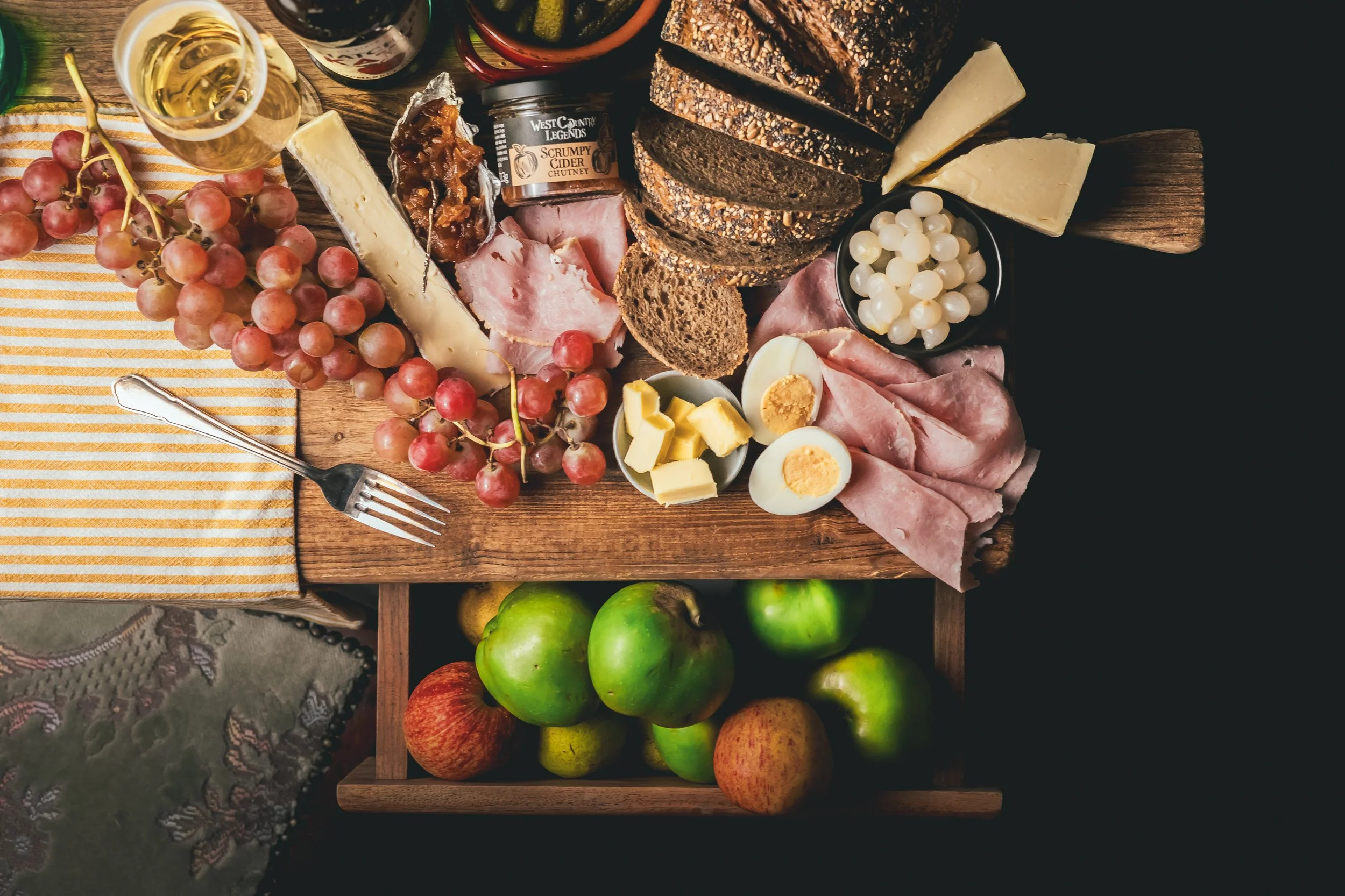 A cheese and charcuterie board with grapes, apples, assorted cheeses, sliced bread, meats, a boiled egg, relish, and a glass of white wine on a wooden table.