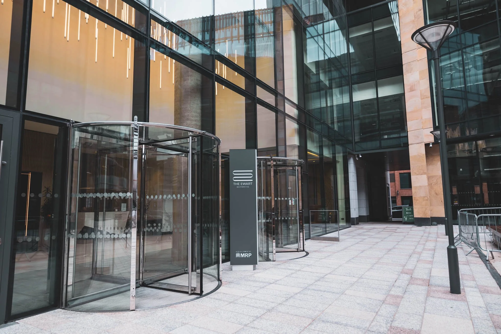 Exterior photography of a modern office building with glass facade, revolving door, and a sign reading 'The Ewart' outside.