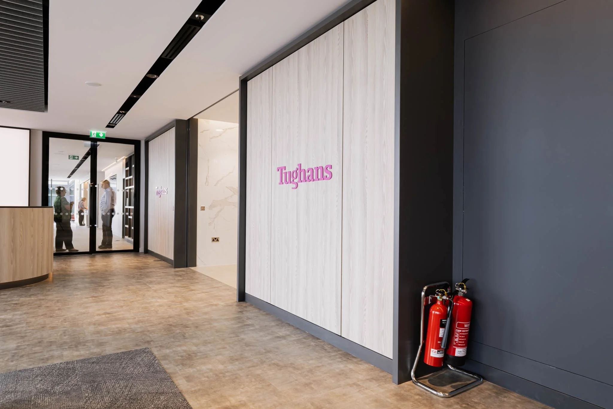 Modern office or lobby entrance with a white wall featuring a pink sign that reads 'Tughans'. There are two fire extinguishers on the floor nearby, and a glass door with people inside visible in the background.