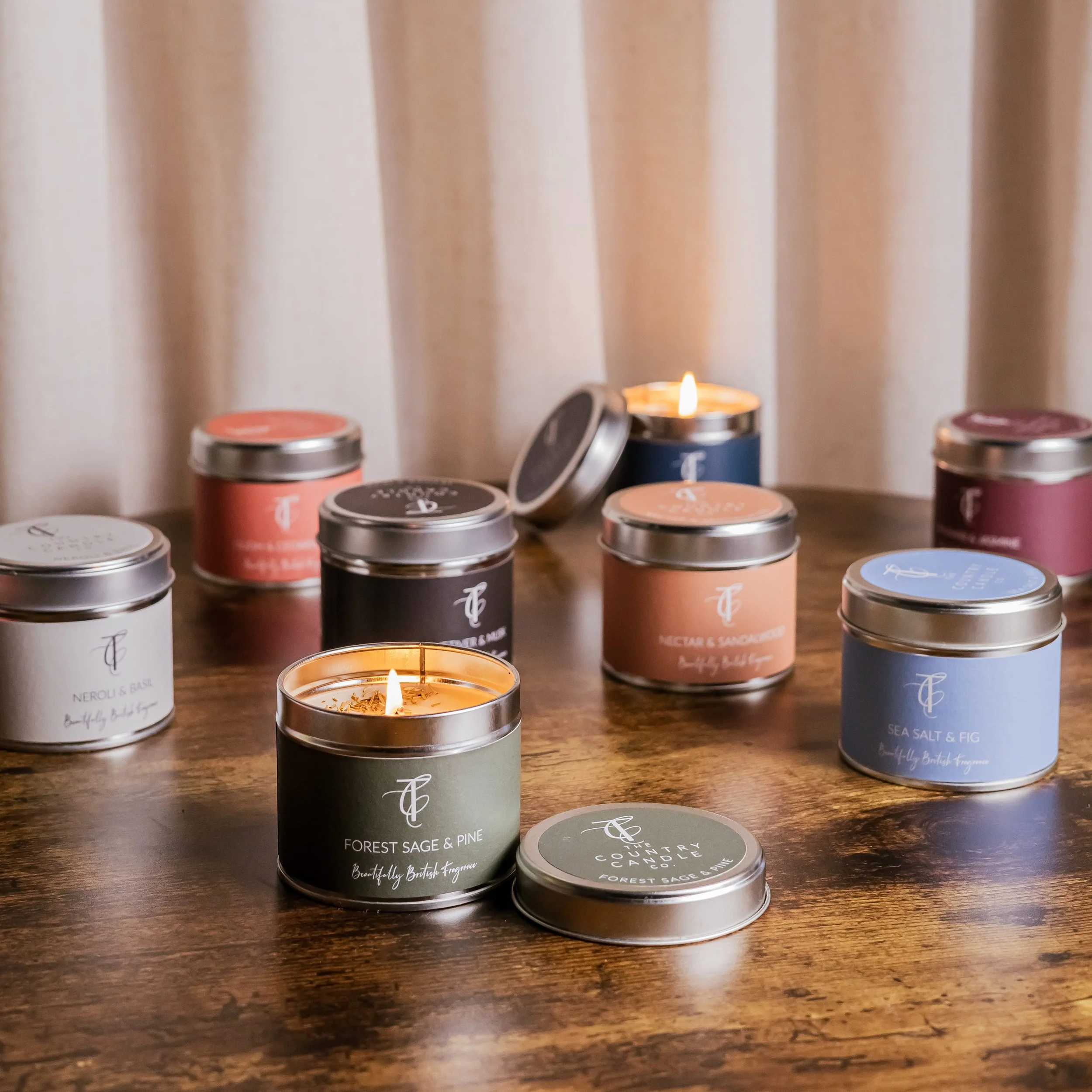 Multiple scented candles in tins with labels, some lit, on a wooden table.