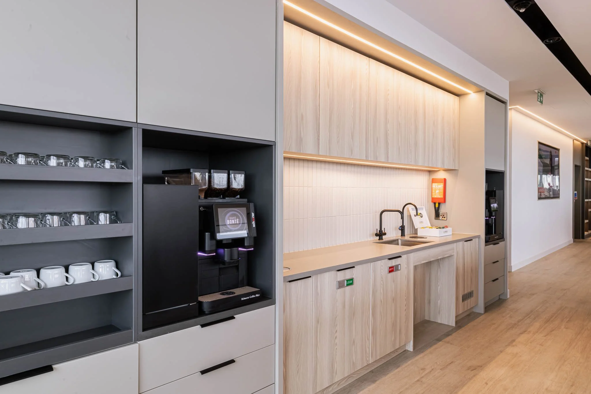 Coffee station with cups, glasses, a coffee machine, and a sink in a modern office or cafe setting with wooden and white cabinetry captured for architecture photography for architects uk