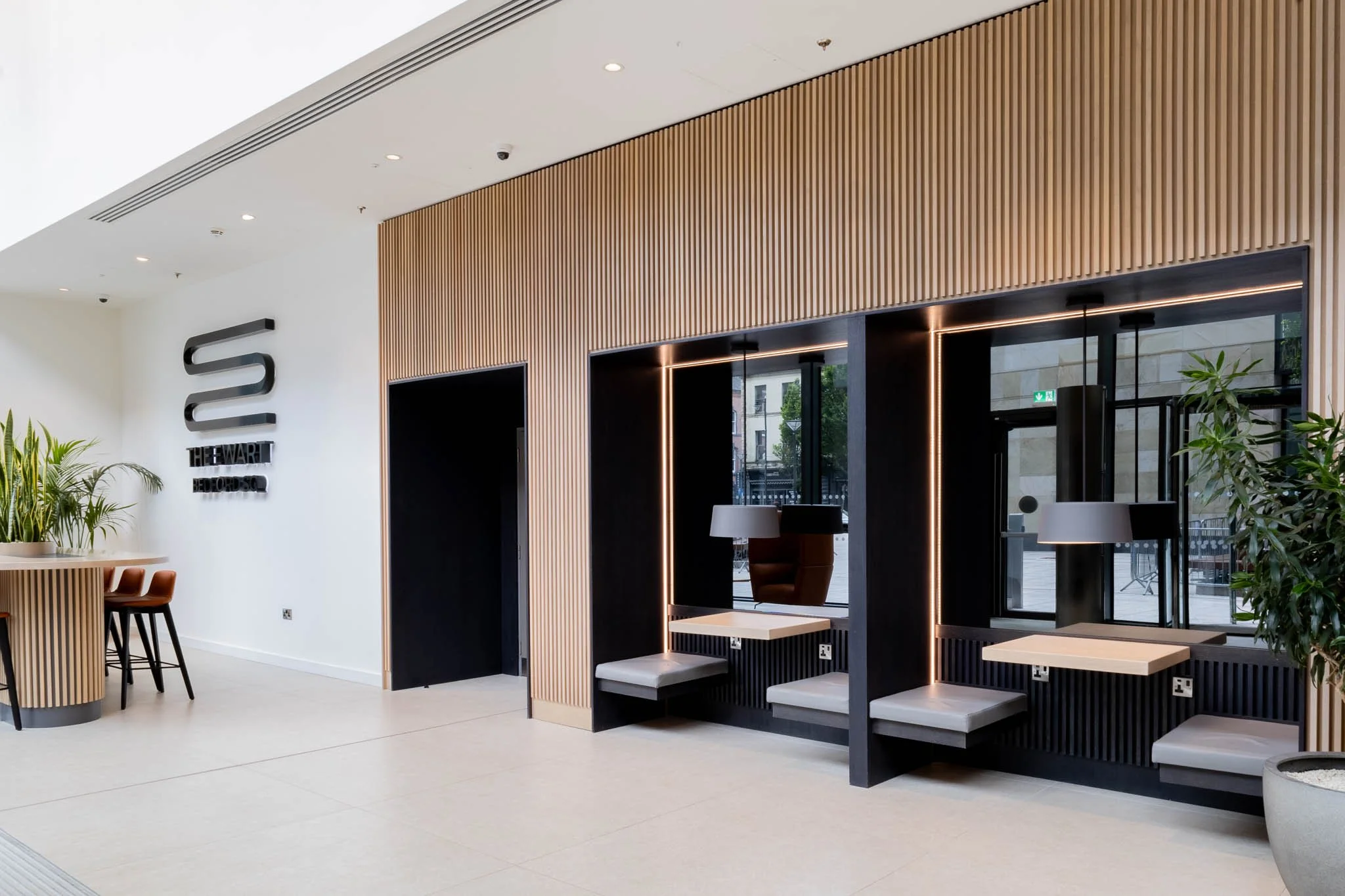 Hotel interior photography UK of modern hotel lobby area with wooden wall, seating booths with cushions, tables, a plant, and a sign that reads 'The Stewart Bedford' on a white wall.