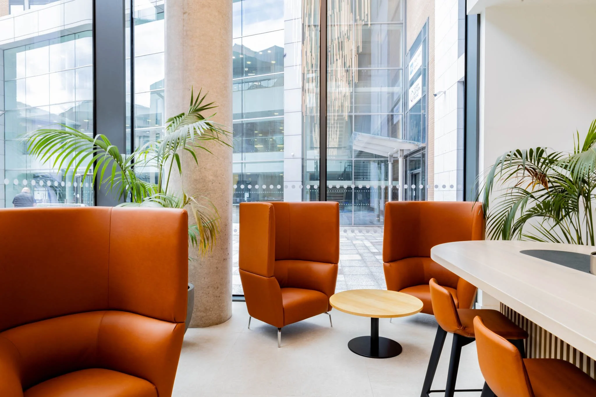 Interior photographer scene of a modern office lobby or lounge area with orange chairs, a small wooden round table, green plants, large floor-to-ceiling windows, and a stone column.