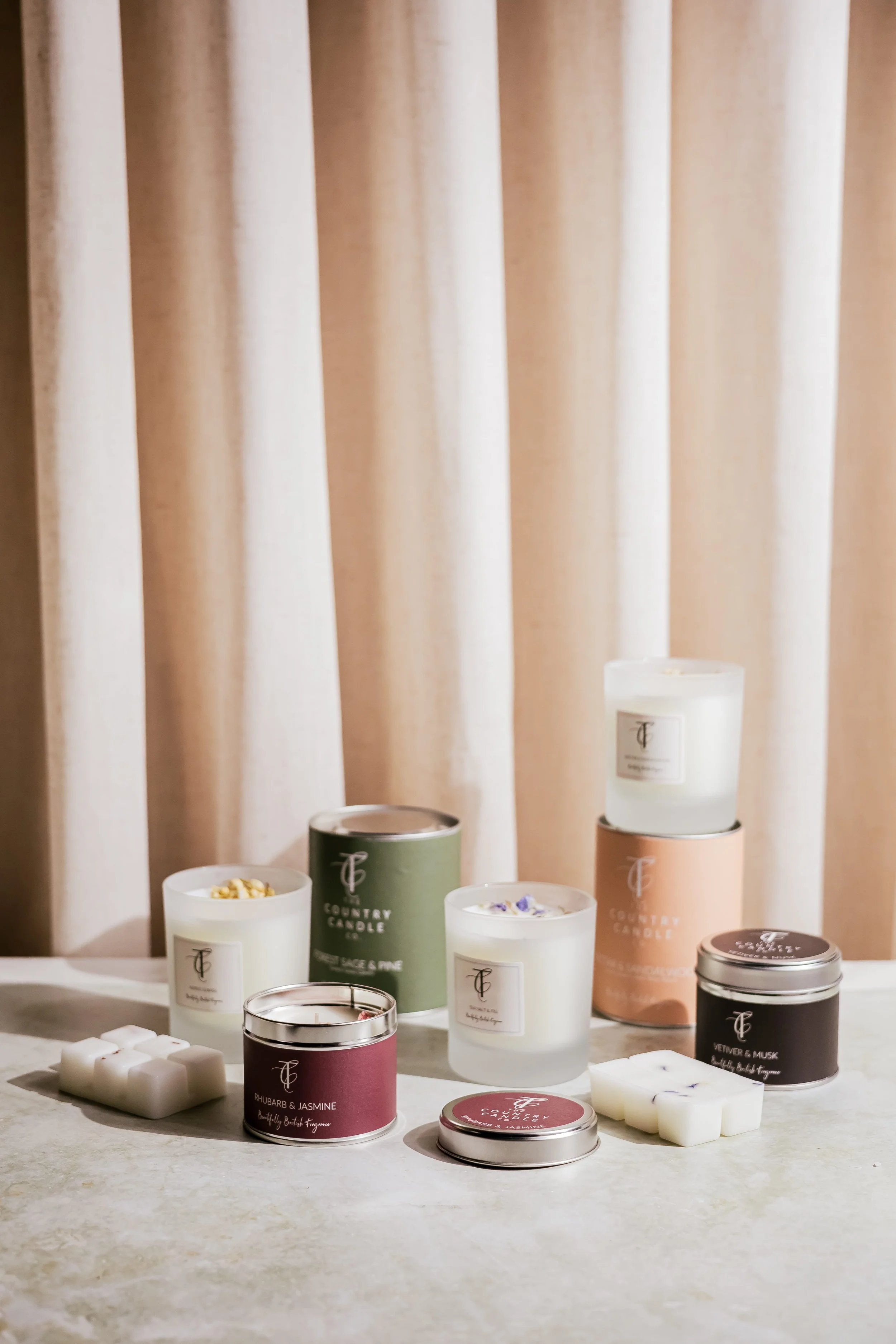A collection of scented candles in various sizes and colors, displayed on a light-colored surface with cream-colored curtains in the background.