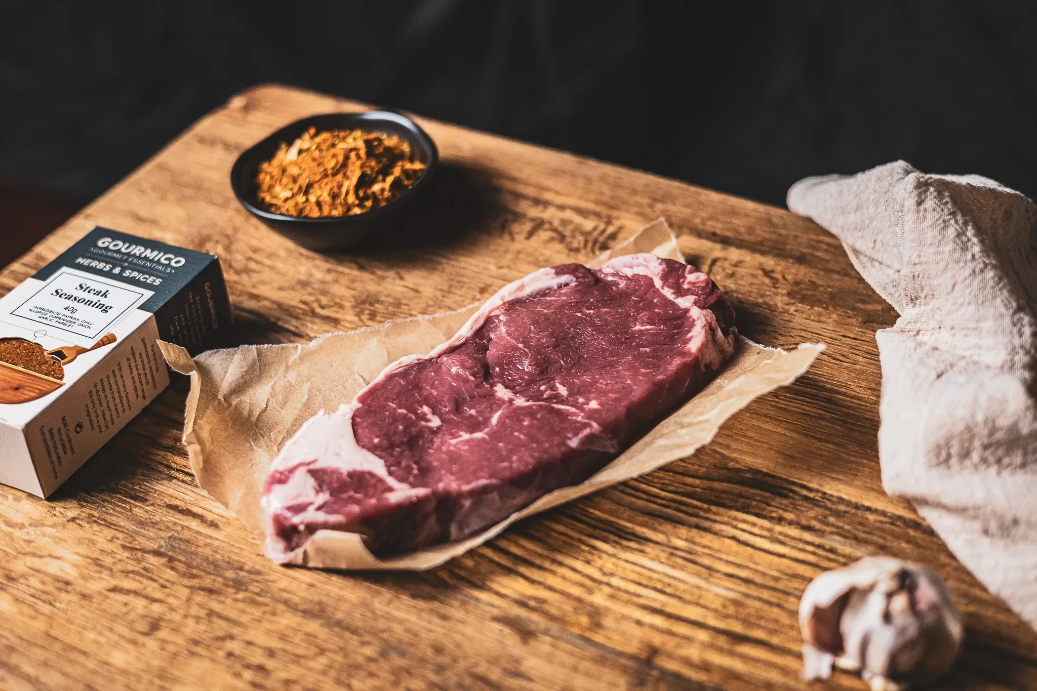 A raw steak on parchment paper, seasoning, garlic, and spices on a wooden cutting board.