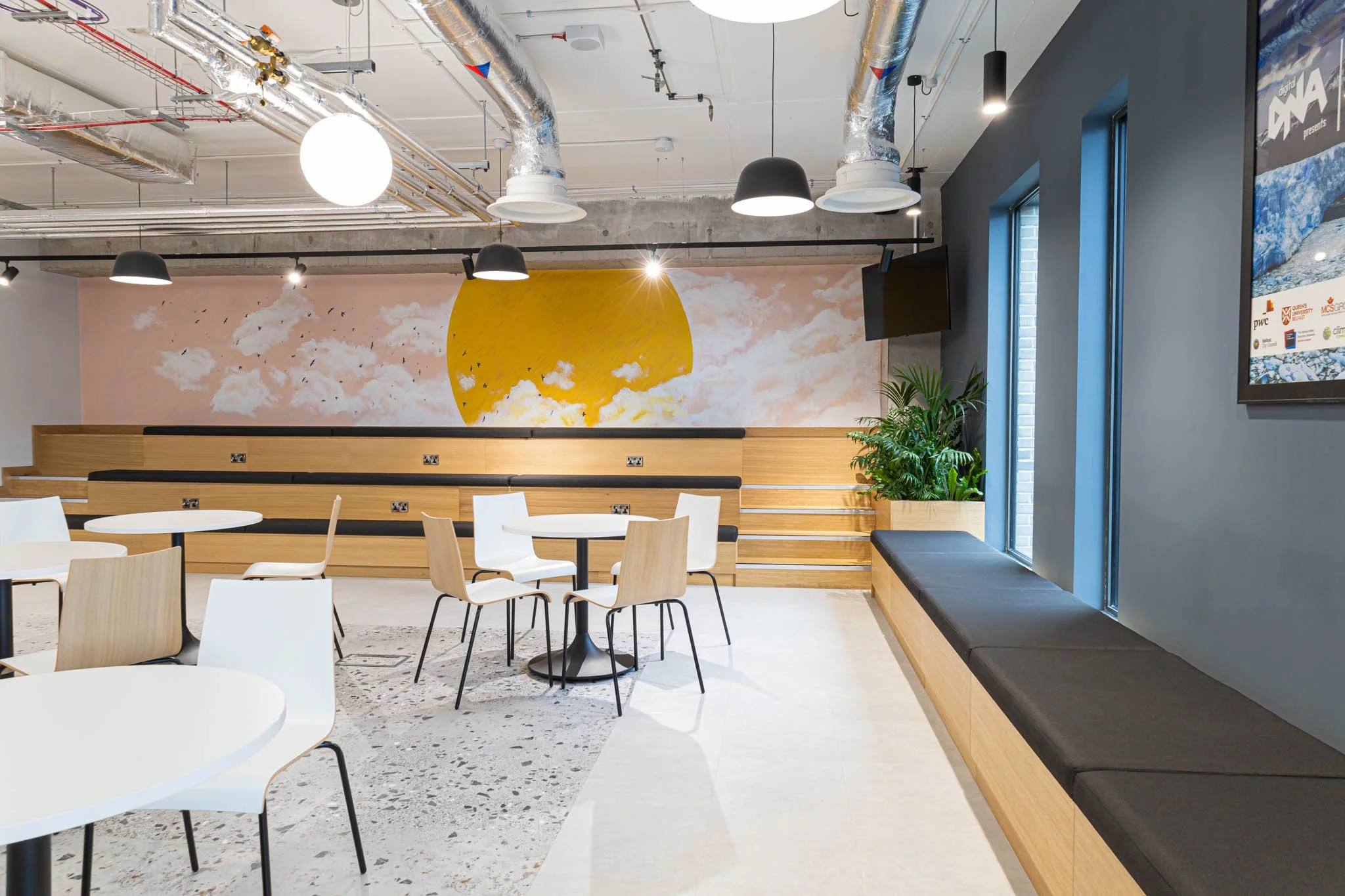 Modern cafe interior photography with wooden benches, white round tables, and chairs, featuring a large mural of a yellow sun and clouds on the wall, black pendant lights, and a window with seating area and potted plants.