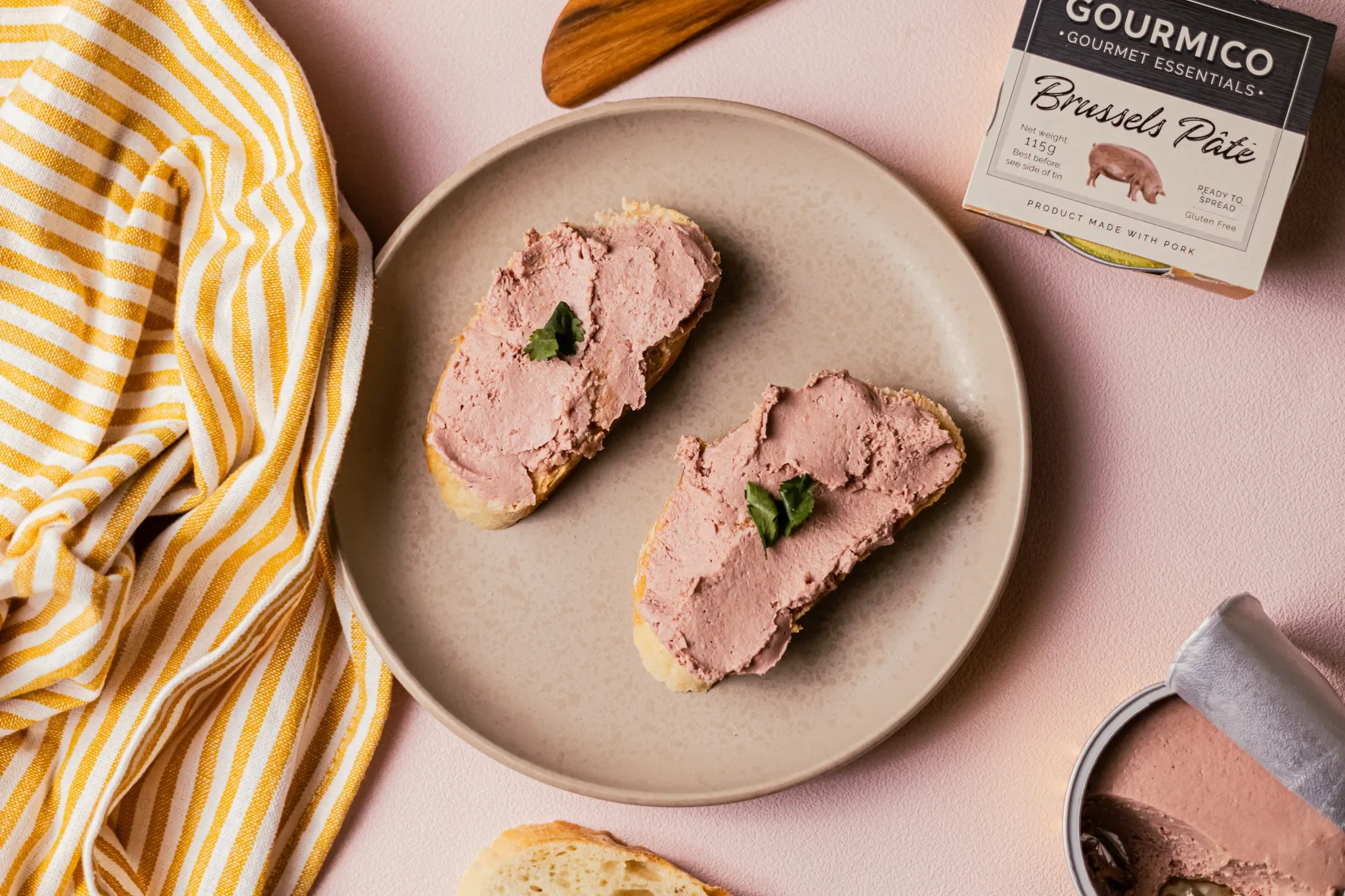 Two pieces of bread with pork liver pâté on a beige plate, garnished with small green herbs, set on a pink surface with a yellow and white striped cloth to the left and a container of pâté to the right.