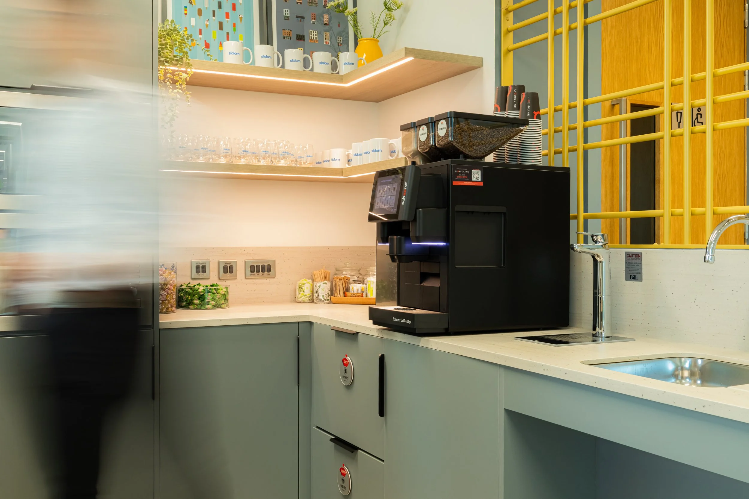 Estate agent photographer services - A modern coffee station with a black coffee machine, cups, and a water faucet on a white countertop. Shelves above hold cups, glasses, and a yellow vase with green plants in Dublin