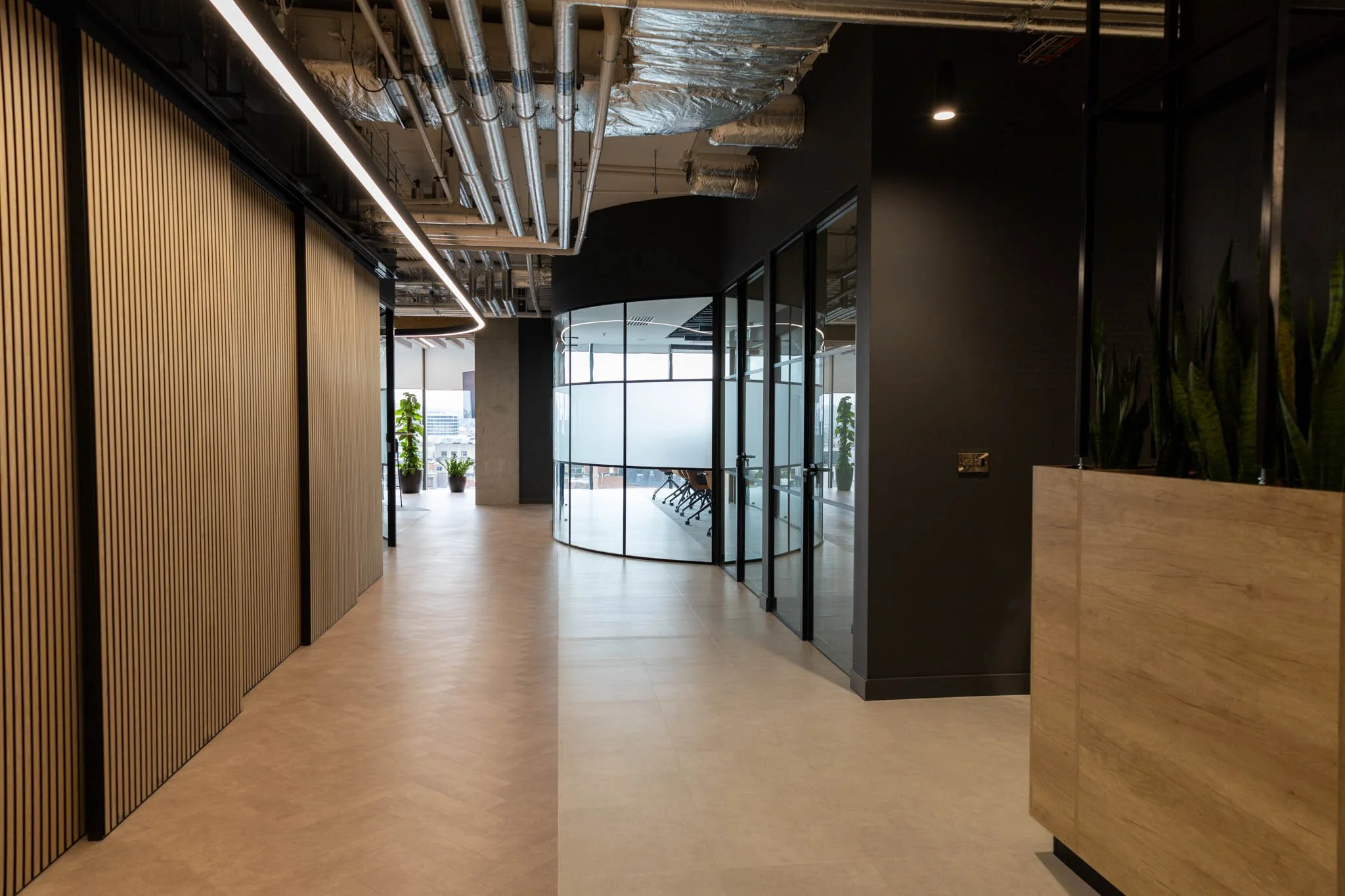 Modern office corridor with wooden slat wall, glass meeting rooms, and potted plants, illuminated by linear ceiling lights.