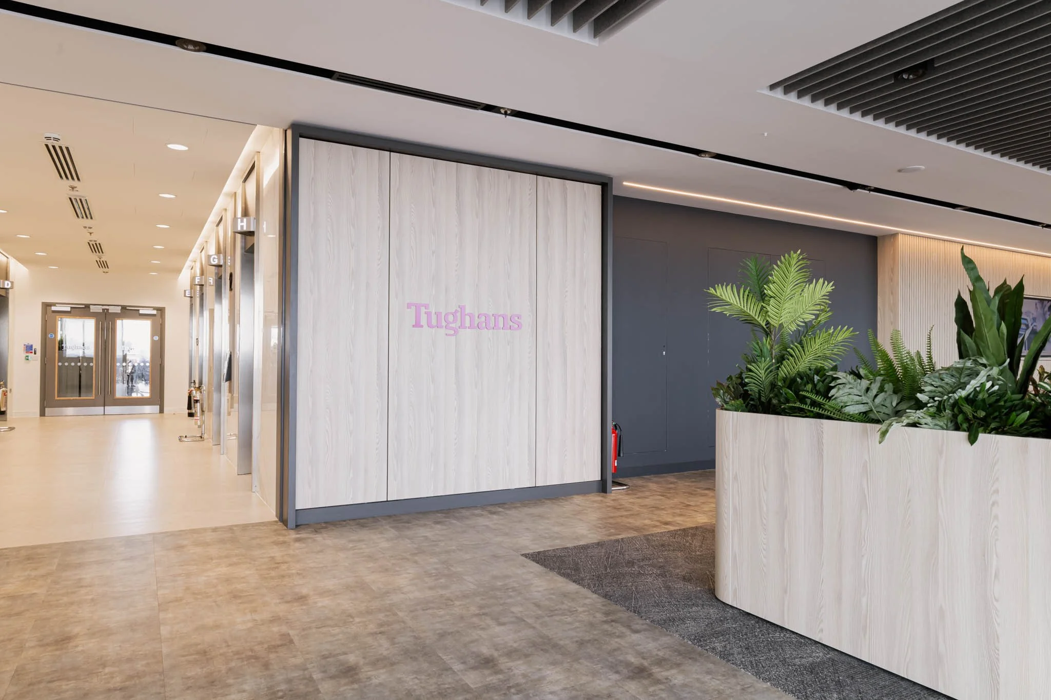 Interior photography of a modern office building lobby with a wooden wall featuring the word "Tughans" in pink, check-in desk with green plants, glass doors in the background.
