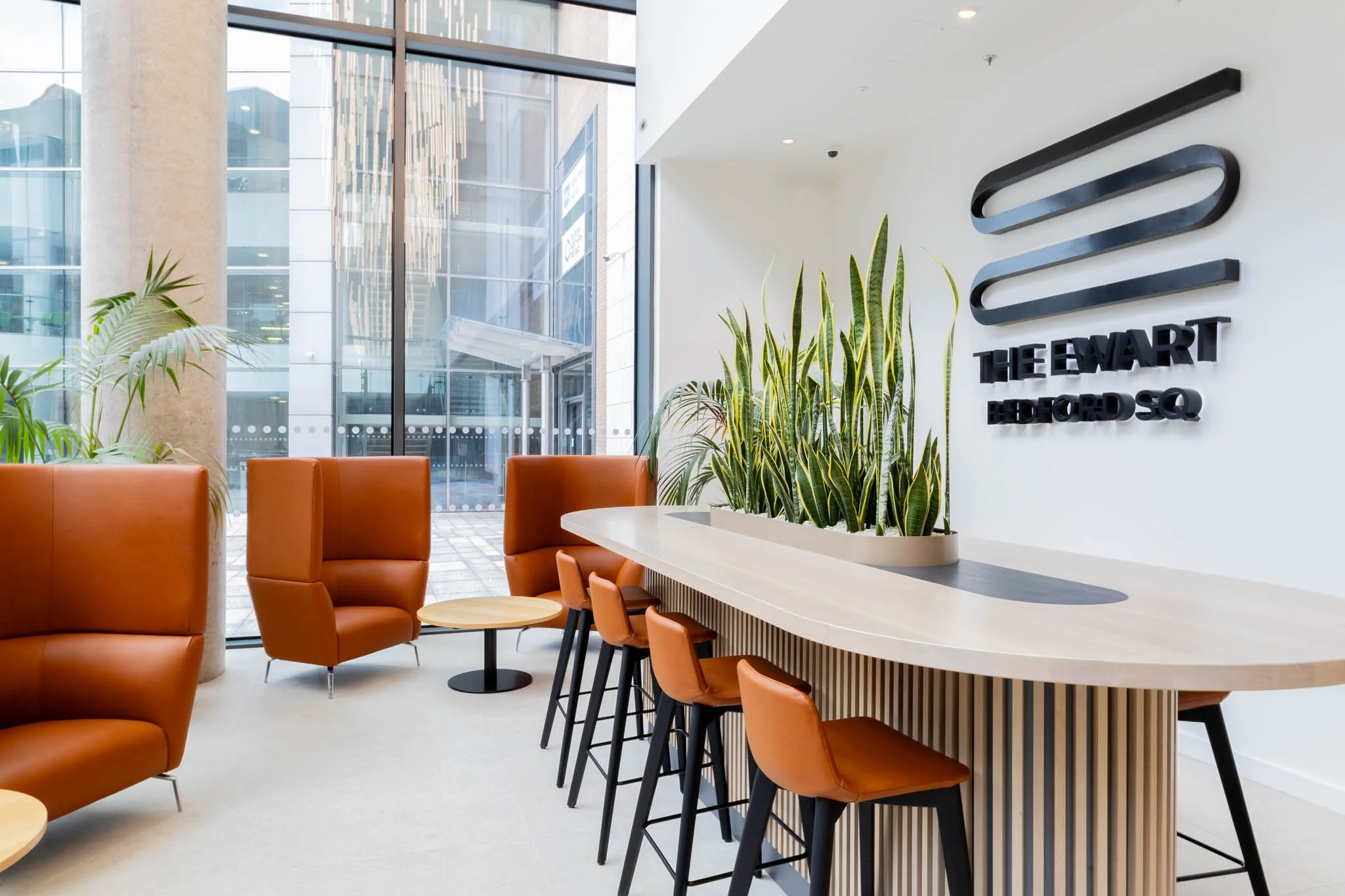 Interior photography of a modern office lobby with leather chairs, a high table with orange chairs, large windows, indoor plants, and a sign on the wall that reads 'The Ewart Bedford Sq.'