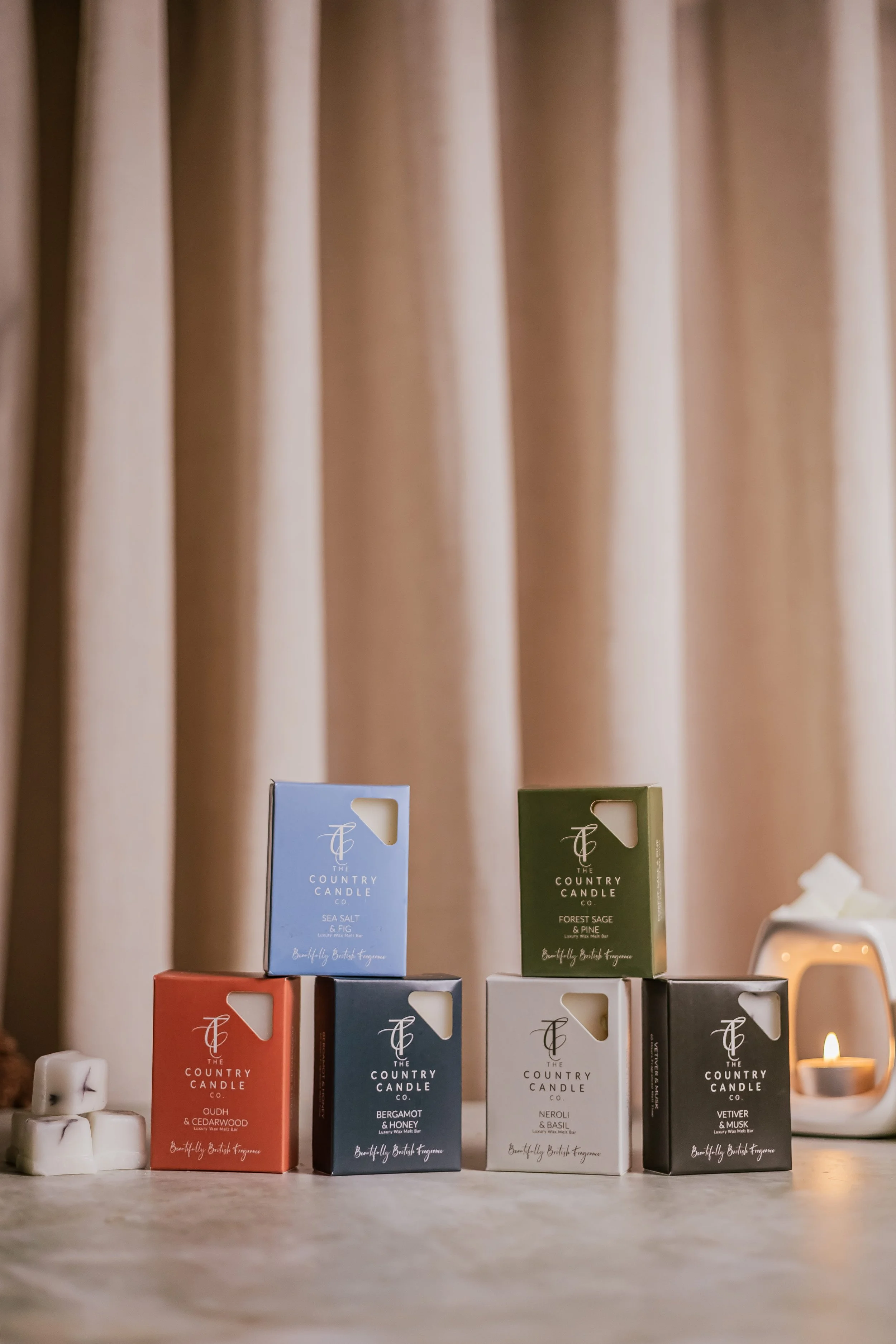 Six boxes of The Country Candle Co. scented candles arranged on a table near a lit candle and some white cubes. The candles are in various colors, matching their scents, with a beige curtain in the background.