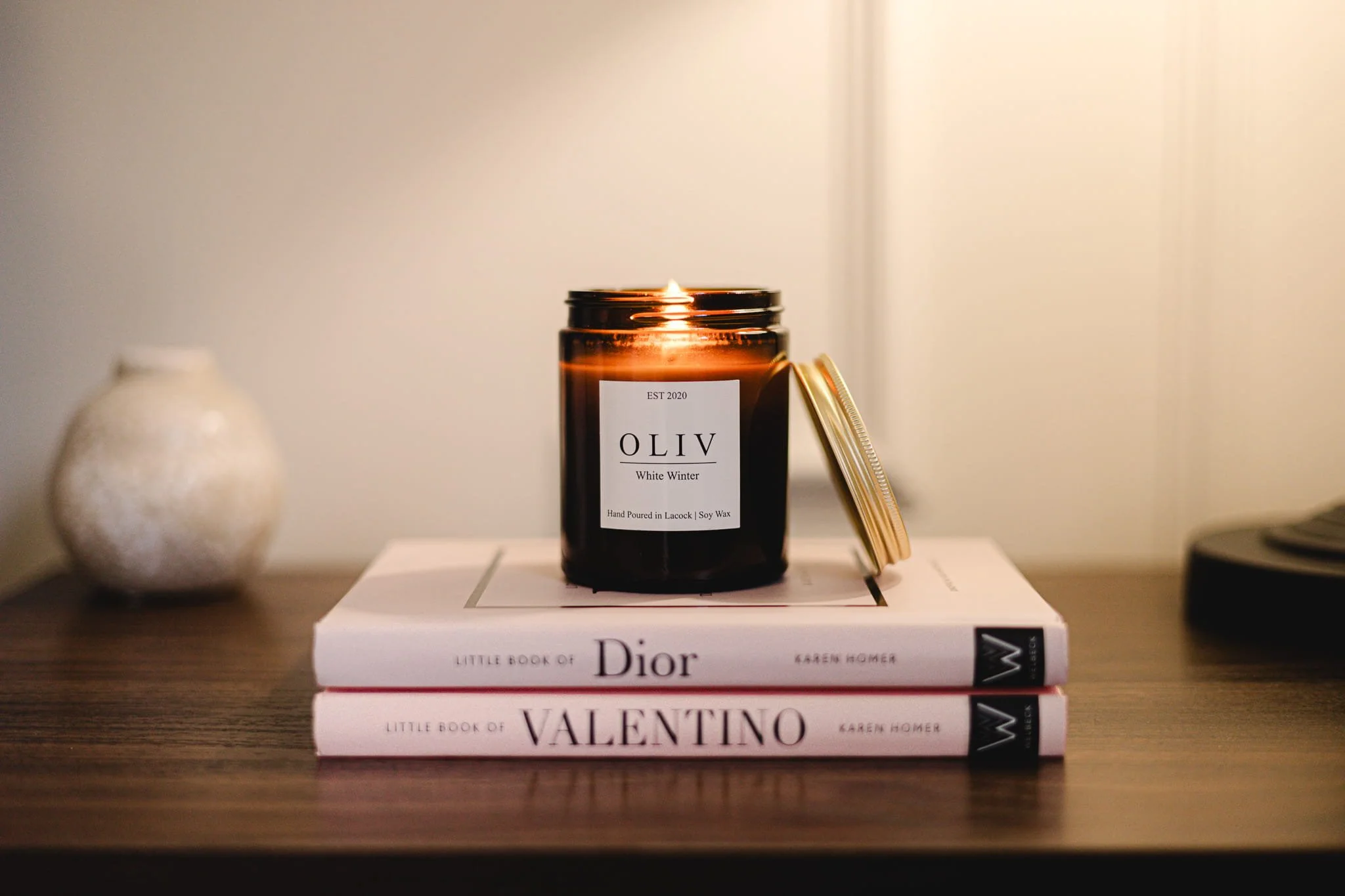 A lit candle in a brown glass jar labeled 'OLIV White Winter,' placed on top of two books titled 'Little Book of Dior' and 'Little Book of Valentino' on a wooden surface.