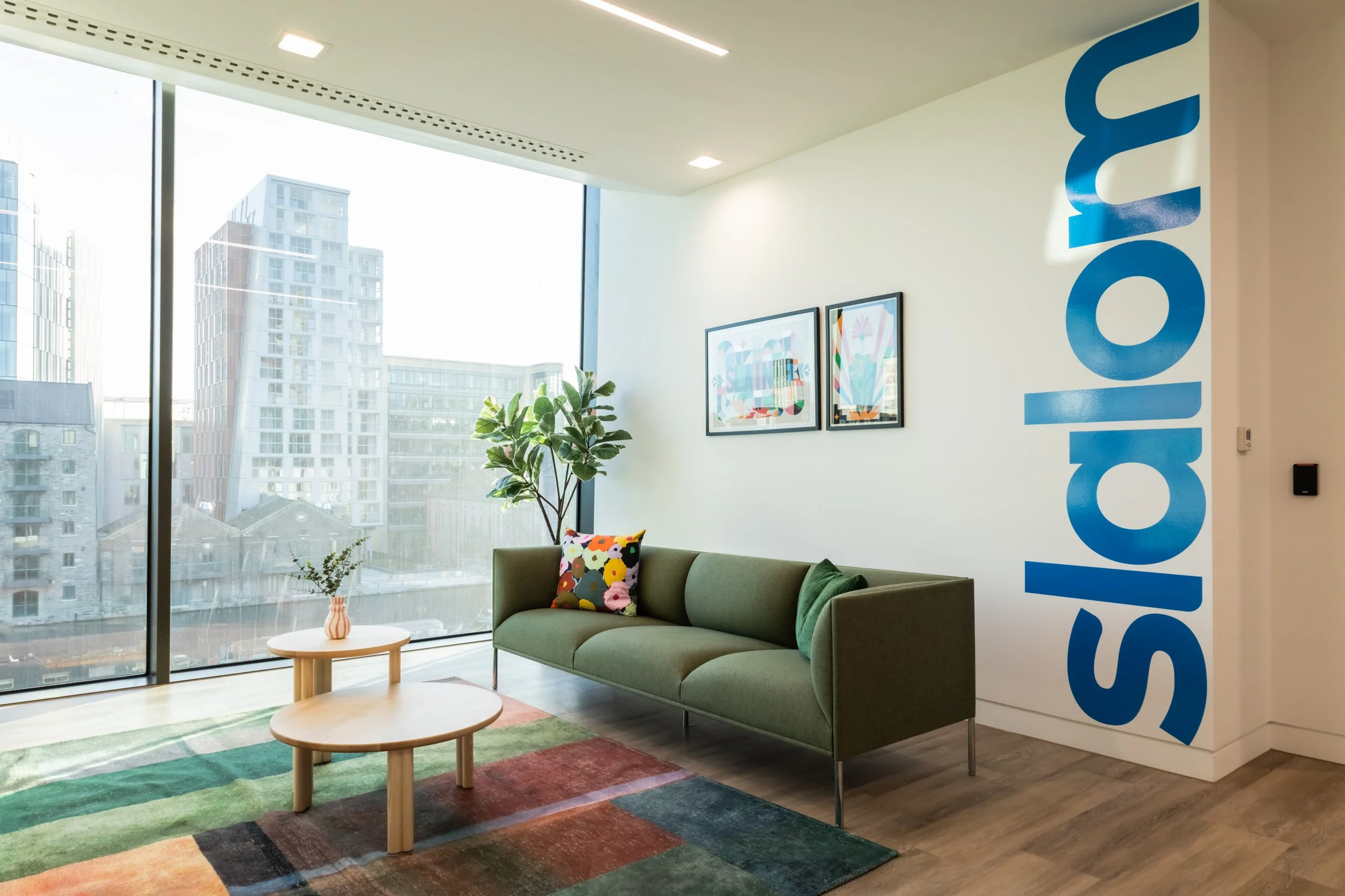 Modern office lounge with green sofa, colorful pillow, two round wooden coffee tables, potted plant, colorful abstract art on white wall, large window with city view, and blue text on wall that reads 'Sydney'.