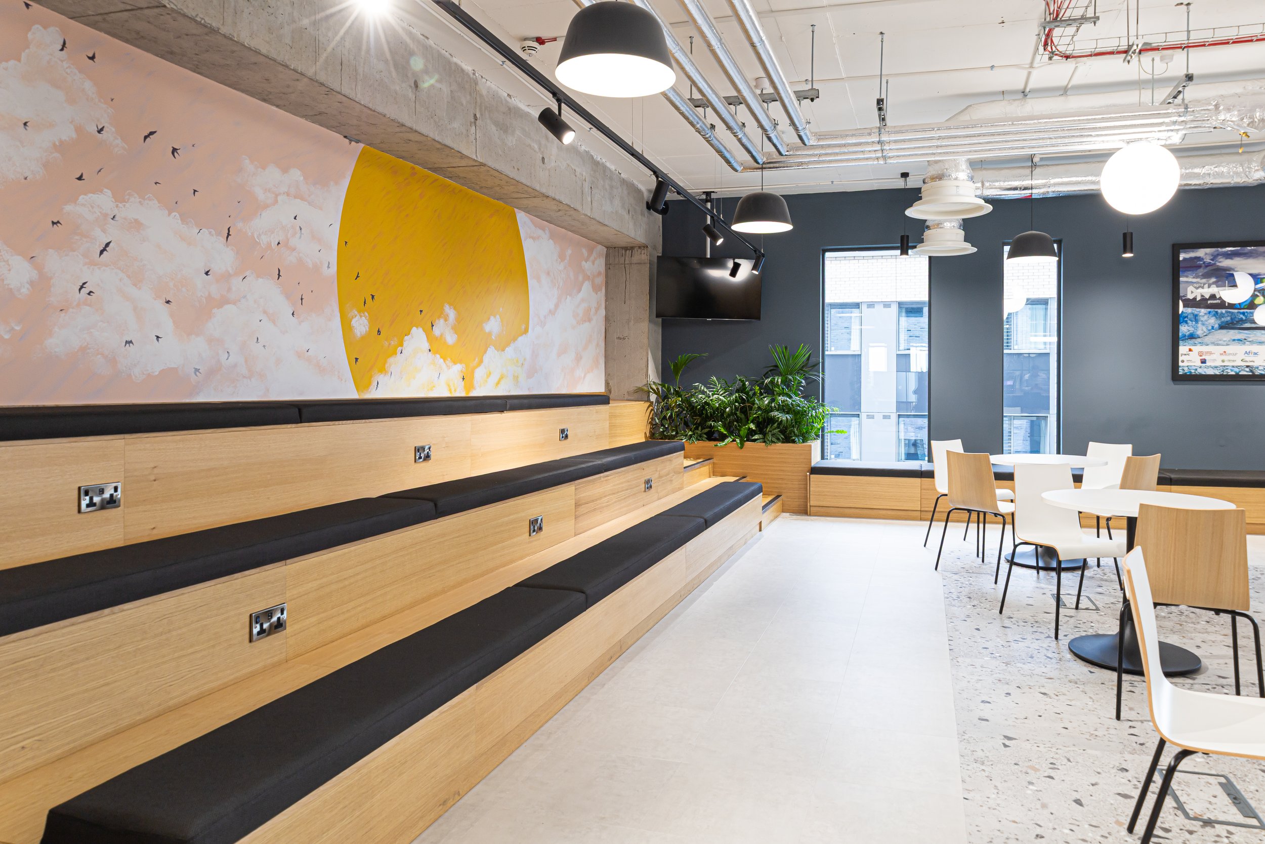 Modern cafe interior with tiered wooden seating, black cushions, white tables, white and wooden chairs, potted plants, large windows, wall artwork of a yellow sun and clouds, ceiling lights, and a blue accent wall.