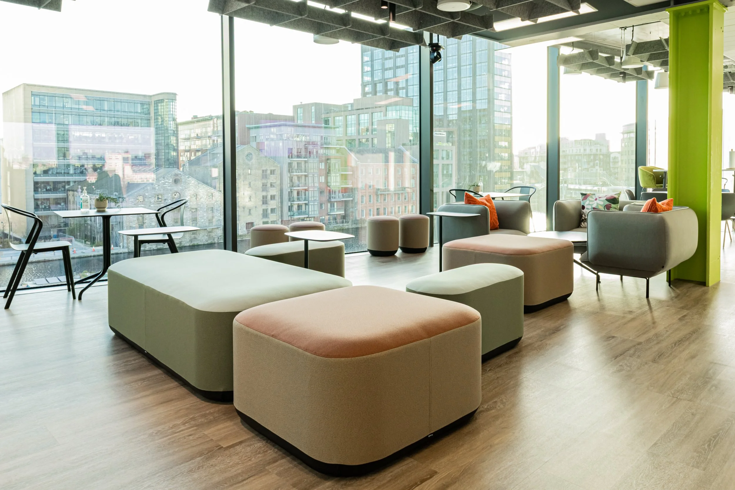 Interior photographer belfast - Modern lounge area with large windows, featuring assorted chairs, sofas, and ottomans in pastel colors, with city buildings visible outside in belfast